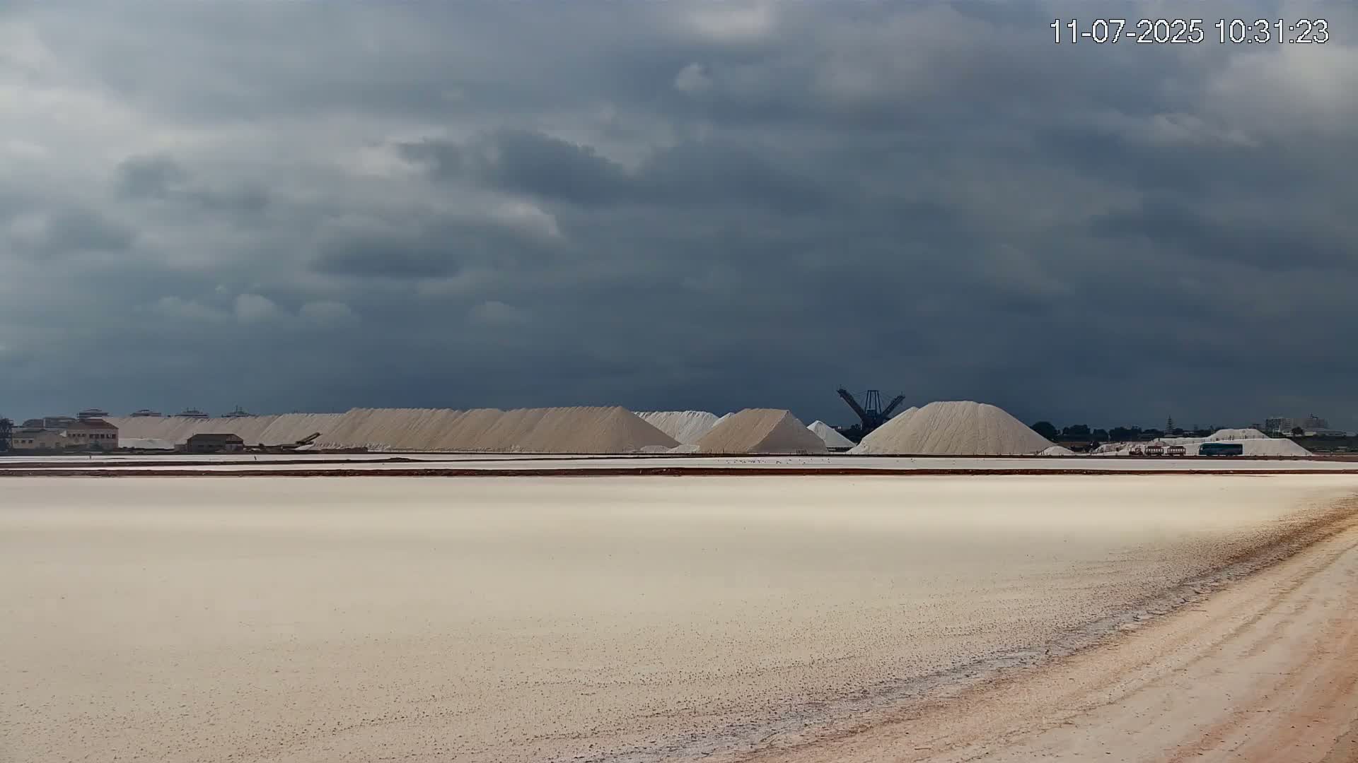 A flat expanse of light beige earth, possibly a salt flat, stretches to a horizon where numerous mounds of white material, likely salt piles, are situated under a cloudy sky.