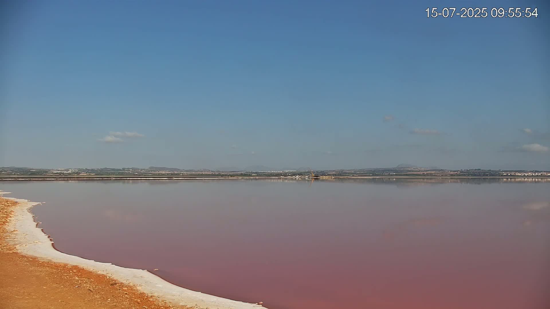 A pink lake stretches to a distant shoreline under a clear blue sky with a few wispy clouds.