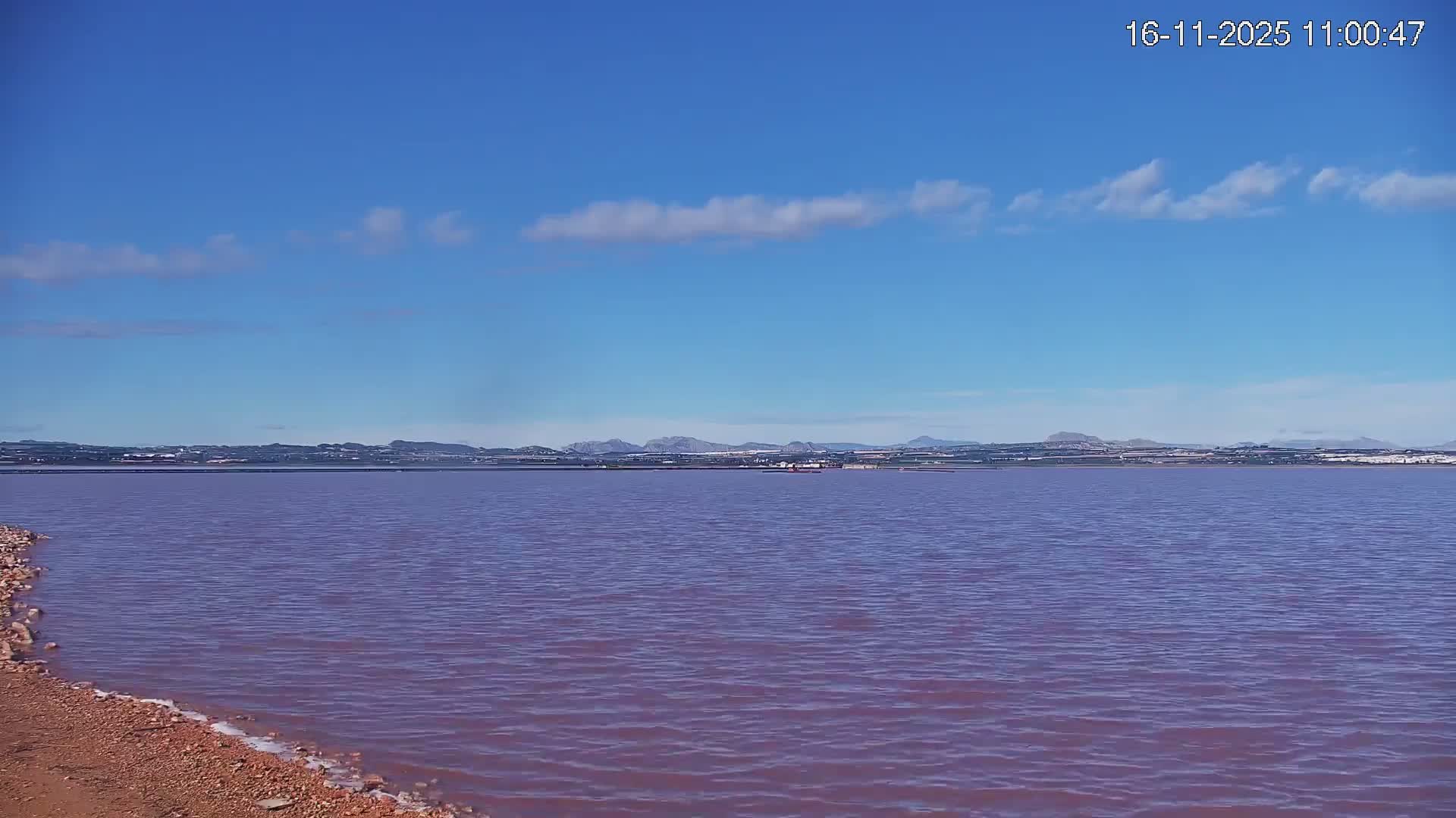 A vibrant pink lake with a reddish rocky shore in the foreground stretches towards distant hills, some with white caps, under a clear blue sky dotted with sparse clouds.
