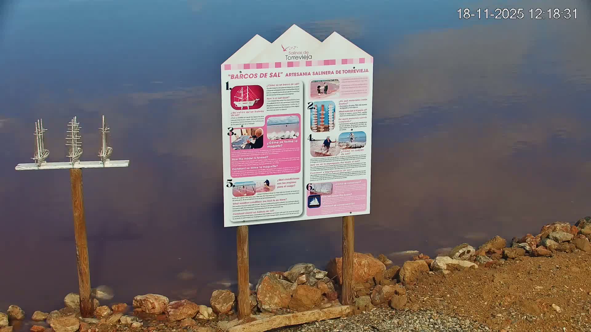 A vibrant pink lake with a reddish rocky shore in the foreground stretches towards distant hills, some with white caps, under a clear blue sky dotted with sparse clouds.