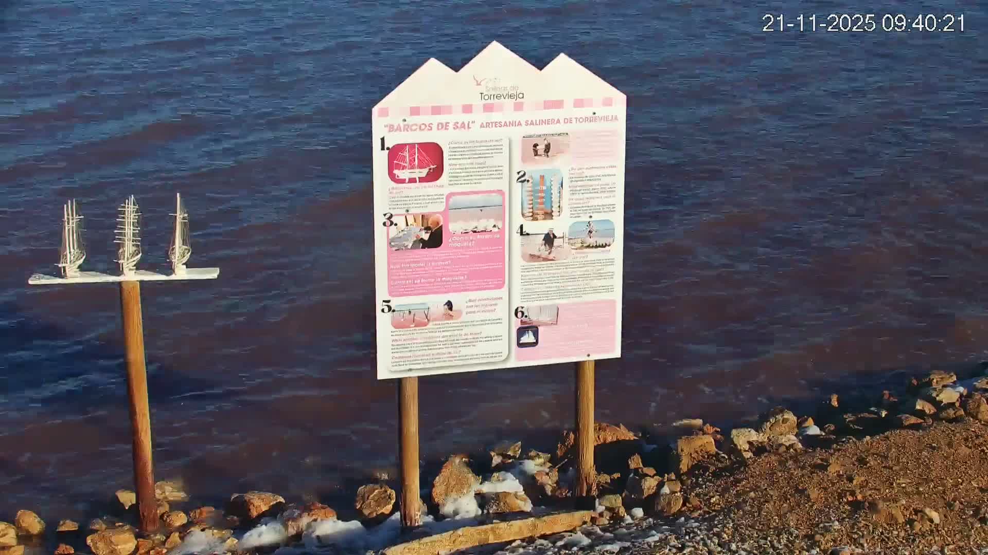 Three miniature sailing ships on a wooden post stand near the rocky, salt-encrusted shore of a large body of water, next to an informational sign, under clear skies.