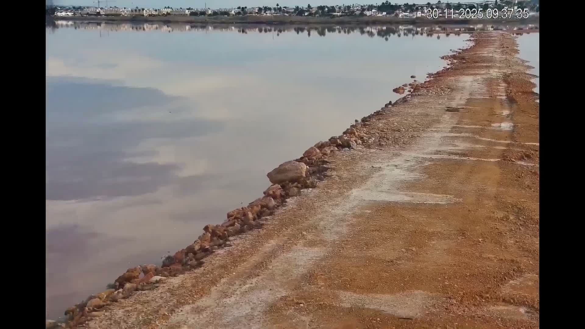 A narrow reddish-brown dirt and rock causeway stretches into a calm, pale body of water reflecting a distant town and a partly cloudy sky, under partly cloudy conditions.