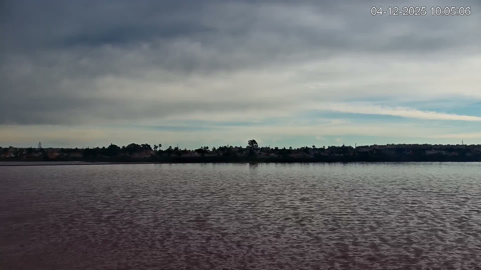 A wide view reveals a vast expanse of rippling, reddish-brown water extending to a distant, dark silhouetted shoreline of a town with trees and structures, all under a heavily overcast sky broken by lighter blue and white cloud streaks.
