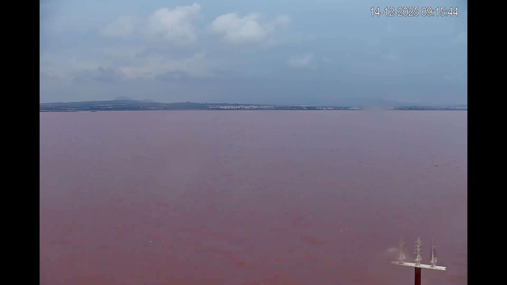 A wide view reveals a vast expanse of rippling, reddish-brown water extending to a distant, dark silhouetted shoreline of a town with trees and structures, all under a heavily overcast sky broken by lighter blue and white cloud streaks.
