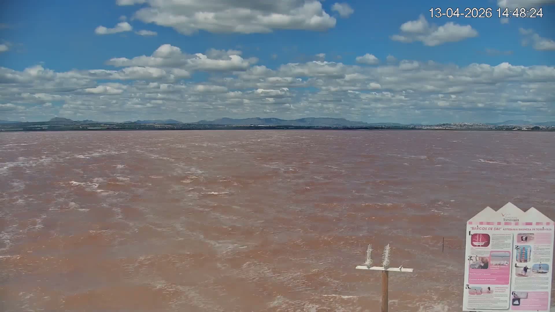 A wide view reveals a vast expanse of rippling, reddish-brown water extending to a distant, dark silhouetted shoreline of a town with trees and structures, all under a heavily overcast sky broken by lighter blue and white cloud streaks.