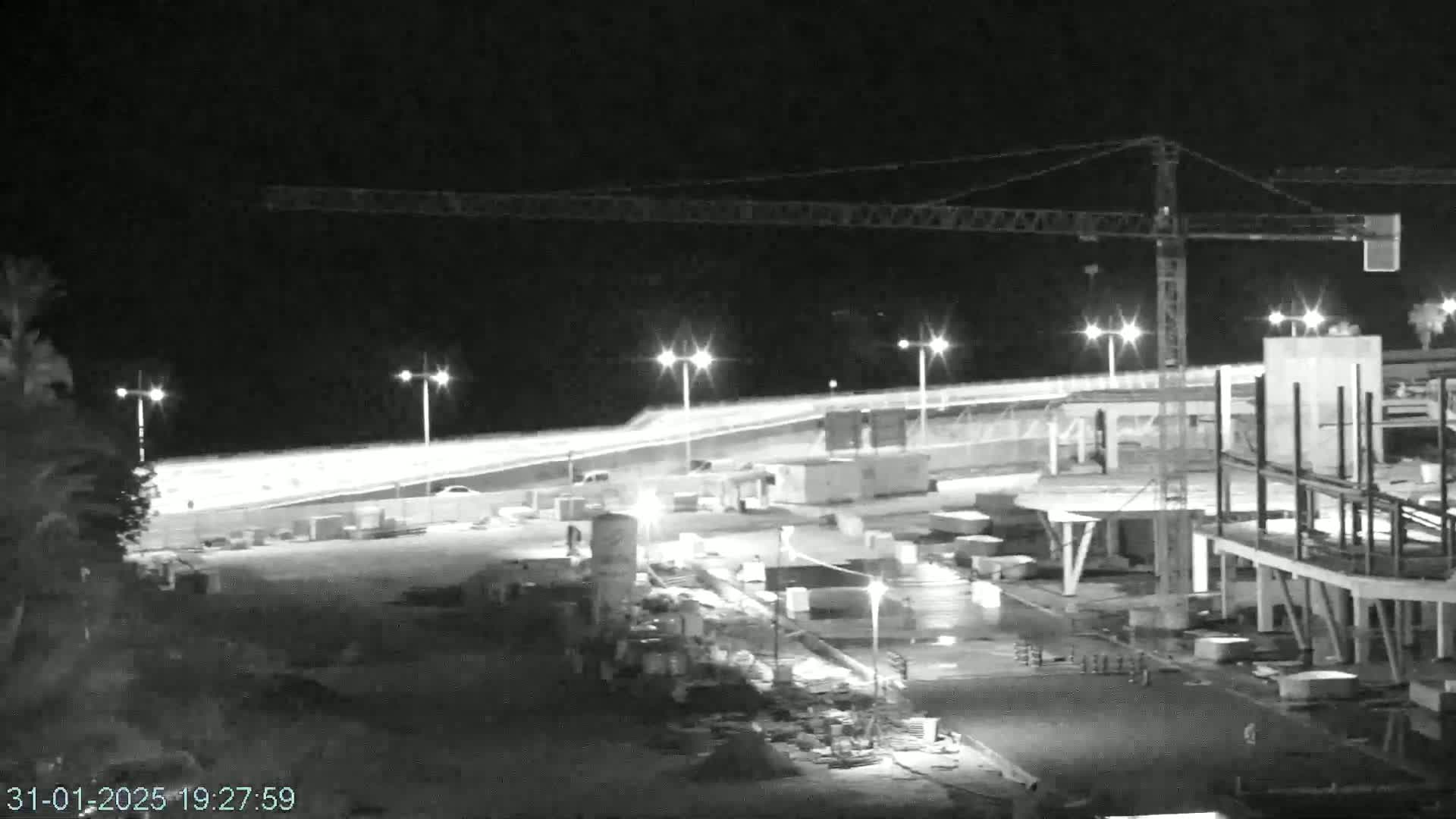 A nighttime, black and white view of a construction site with a crane, floodlights, and a partially built structure.