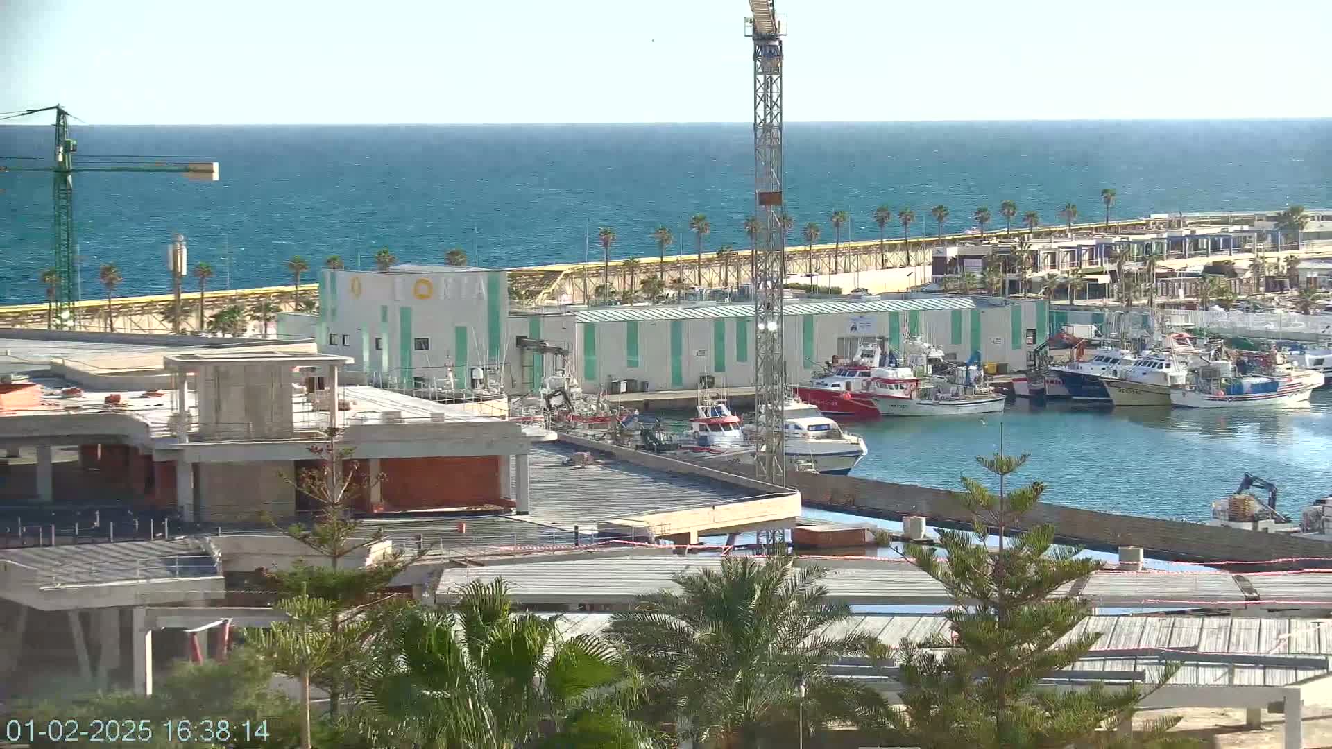 A harbor filled with numerous fishing boats is situated next to a partially constructed building and a calm blue sea under a clear sky.