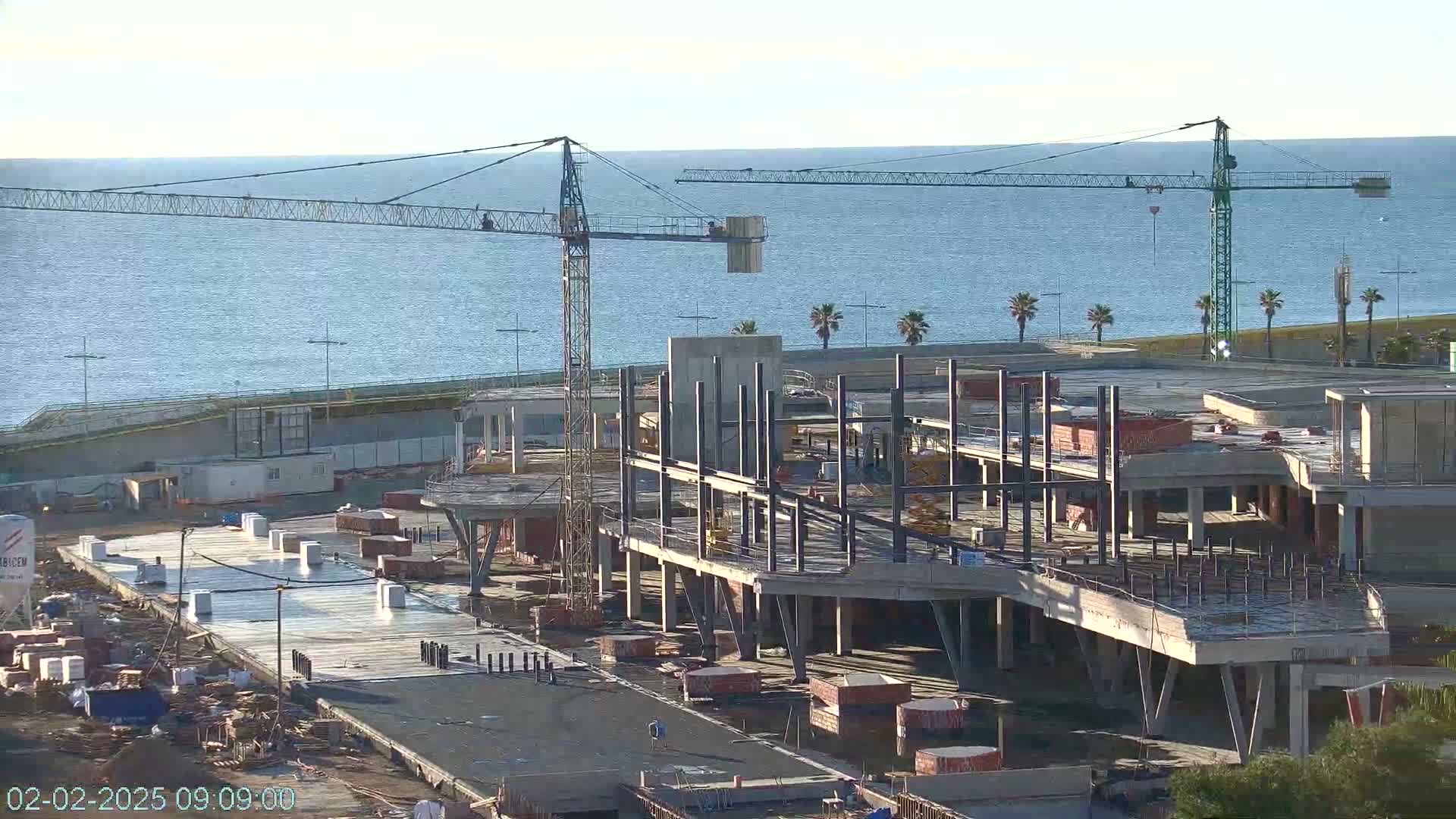 A large construction site with several cranes is visible near a calm body of water under a clear sky.