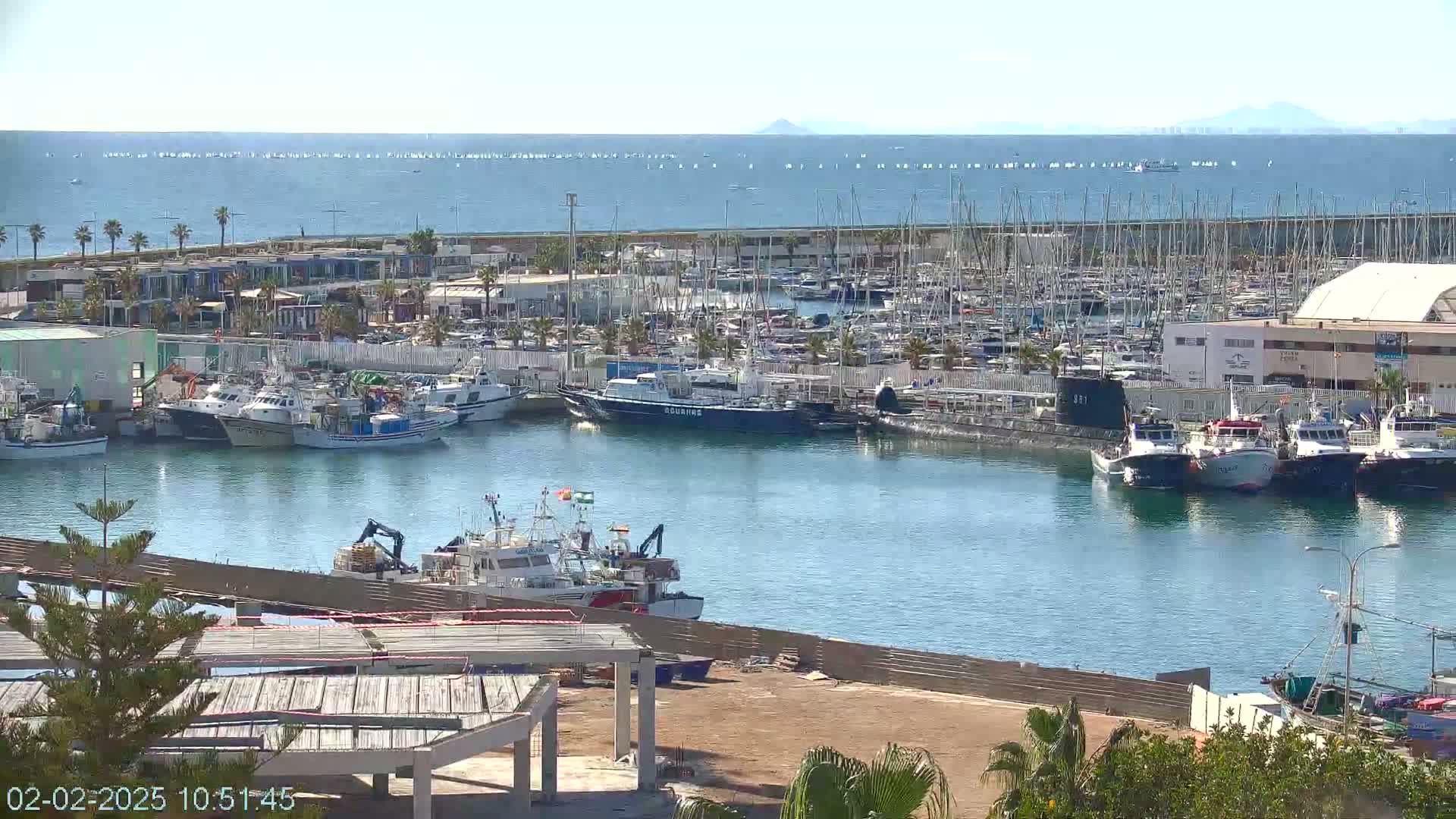 A marina filled with numerous boats and ships, some docked and others at sea, under a clear sunny sky.