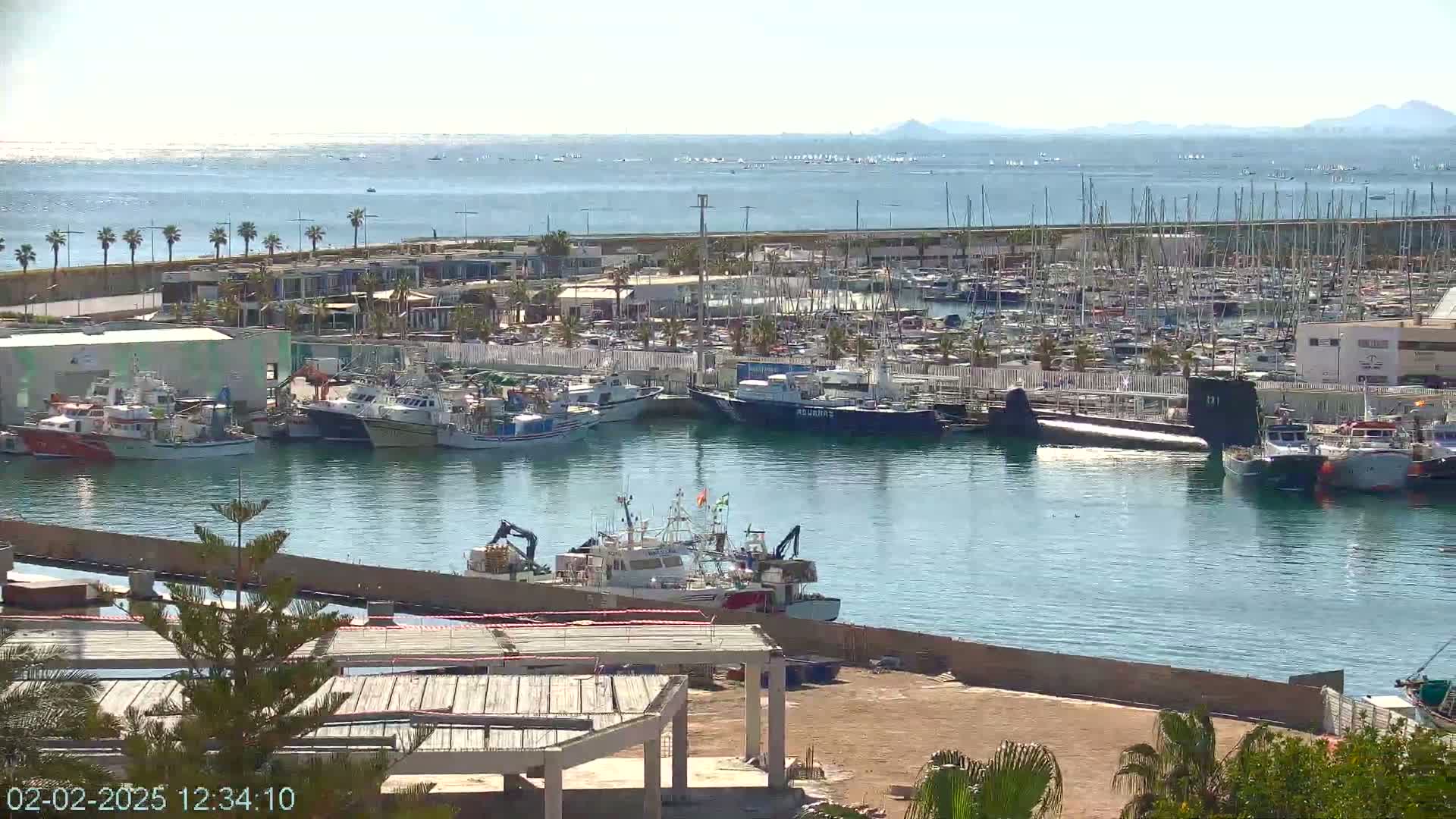 A harbor filled with numerous boats and a submarine, under sunny skies, with a calm sea extending to distant mountains in the background.