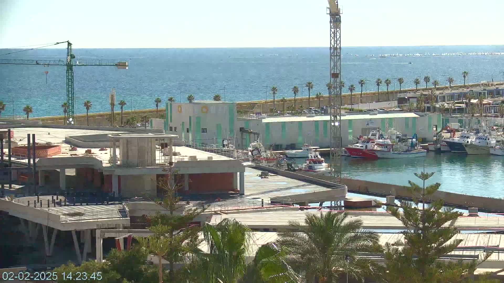 A partly constructed building overlooks a marina filled with boats on a calm, sunny day next to a blue ocean.