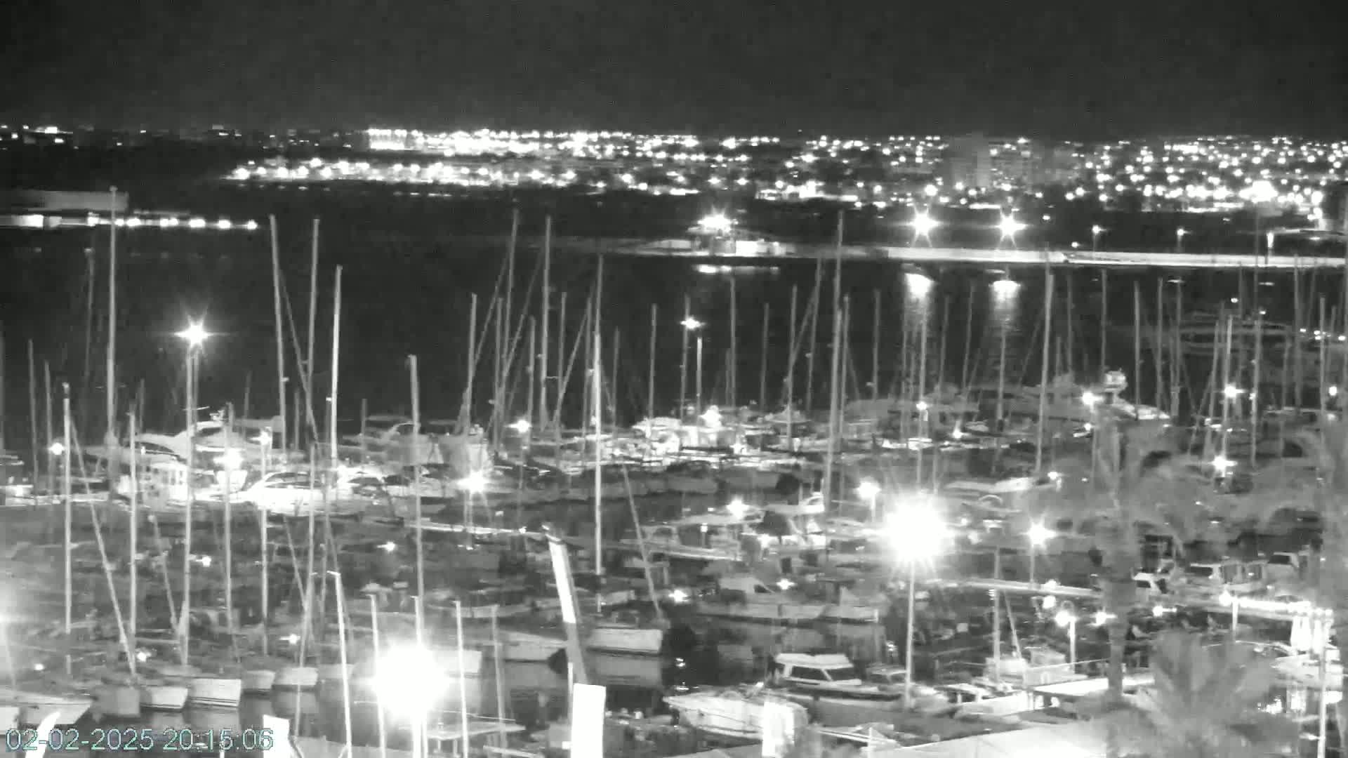 A nighttime, high-angle view of a marina filled with numerous boats and yachts, illuminated by lights, with a city skyline visible in the background across the water.
