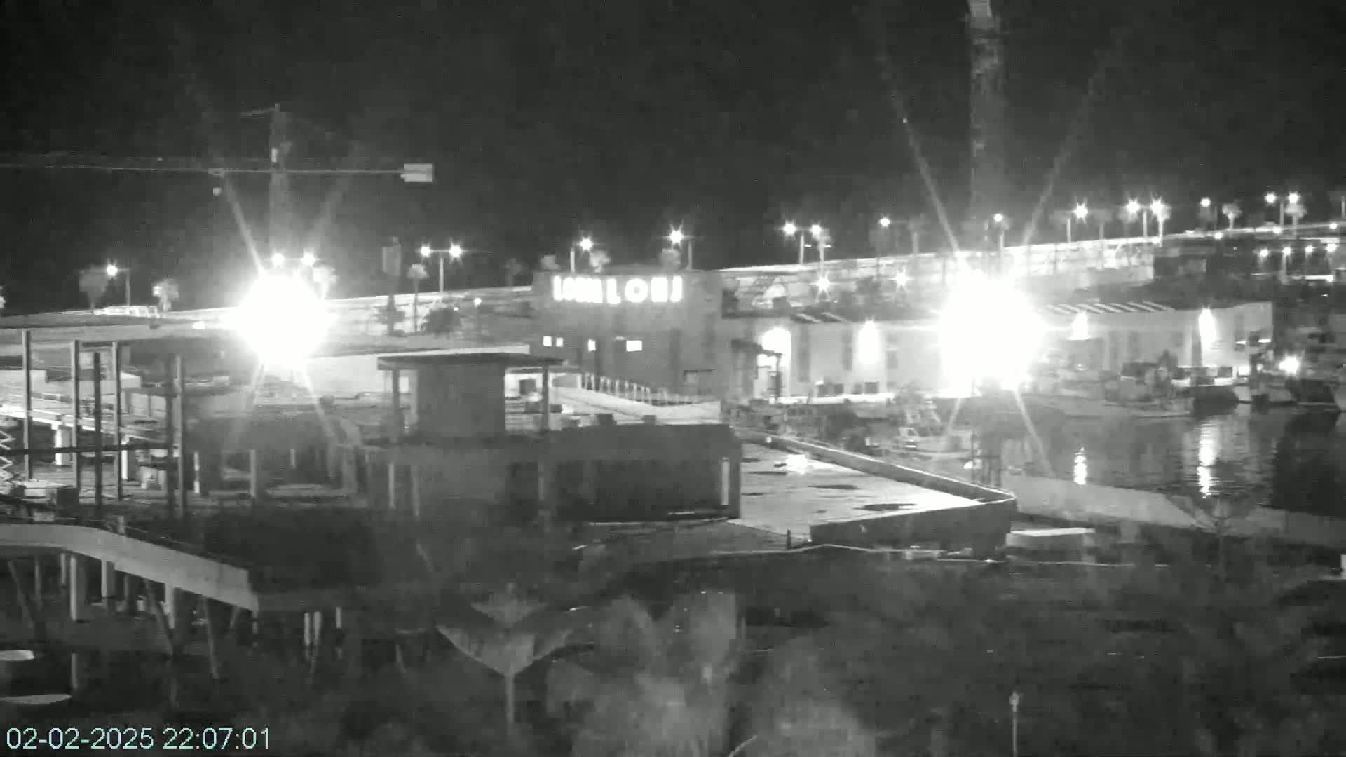 A nighttime, low-light view shows a partially constructed building near a body of water with lights illuminating a dock and boats.