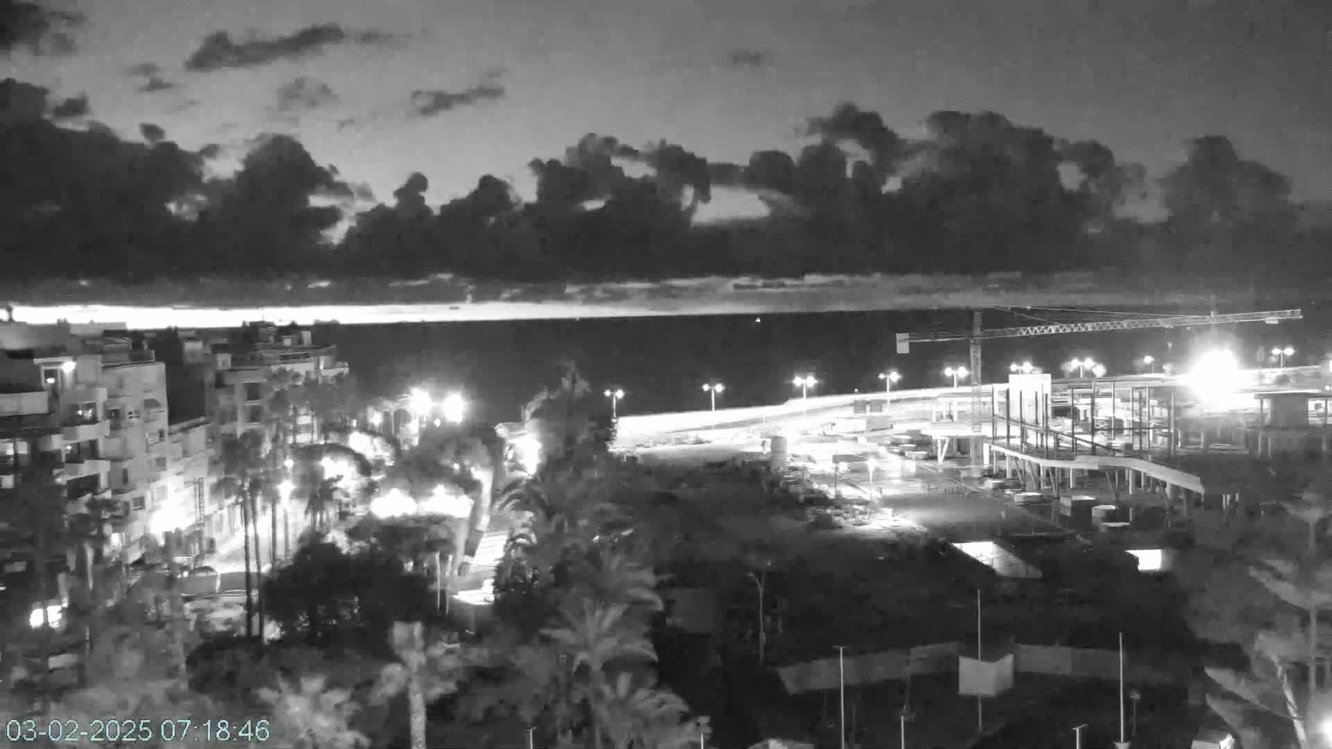 A nighttime, overcast view shows a coastal city with palm trees, buildings, and a construction site near the water.