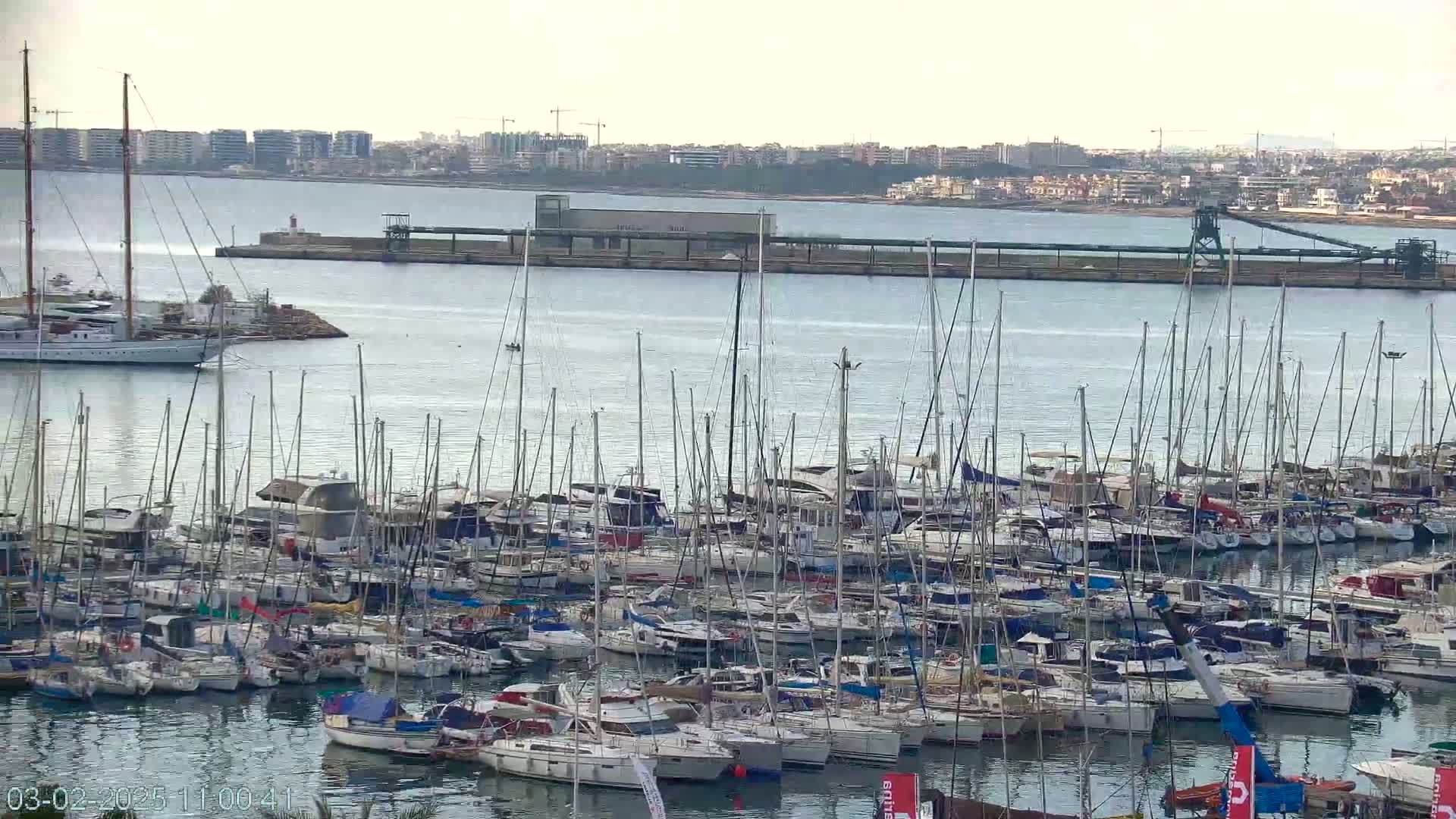 A large marina filled with numerous sailboats and motorboats is situated on calm water under an overcast sky, with a distant cityscape visible across the bay.