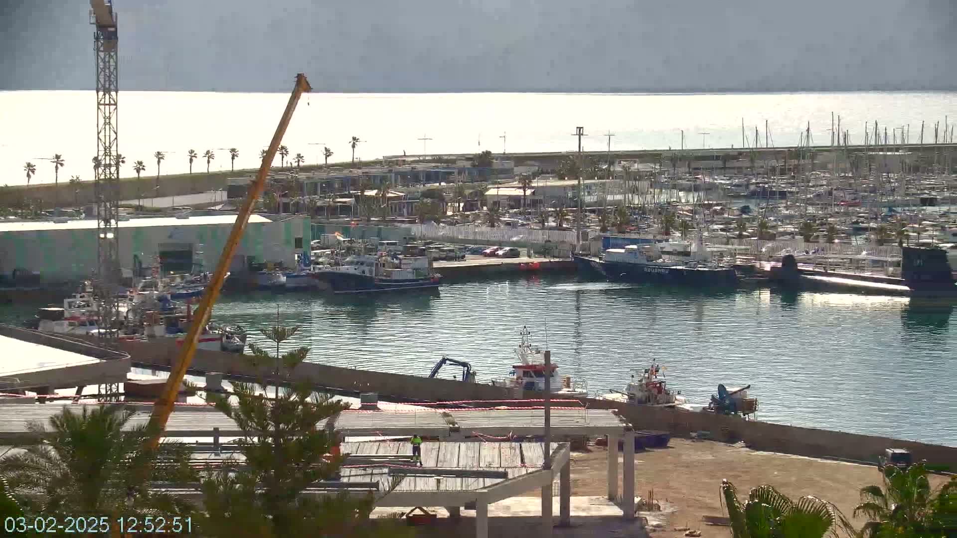 A harbor filled with numerous boats and yachts, under a mostly sunny sky, with a crane and construction underway on the lower right.