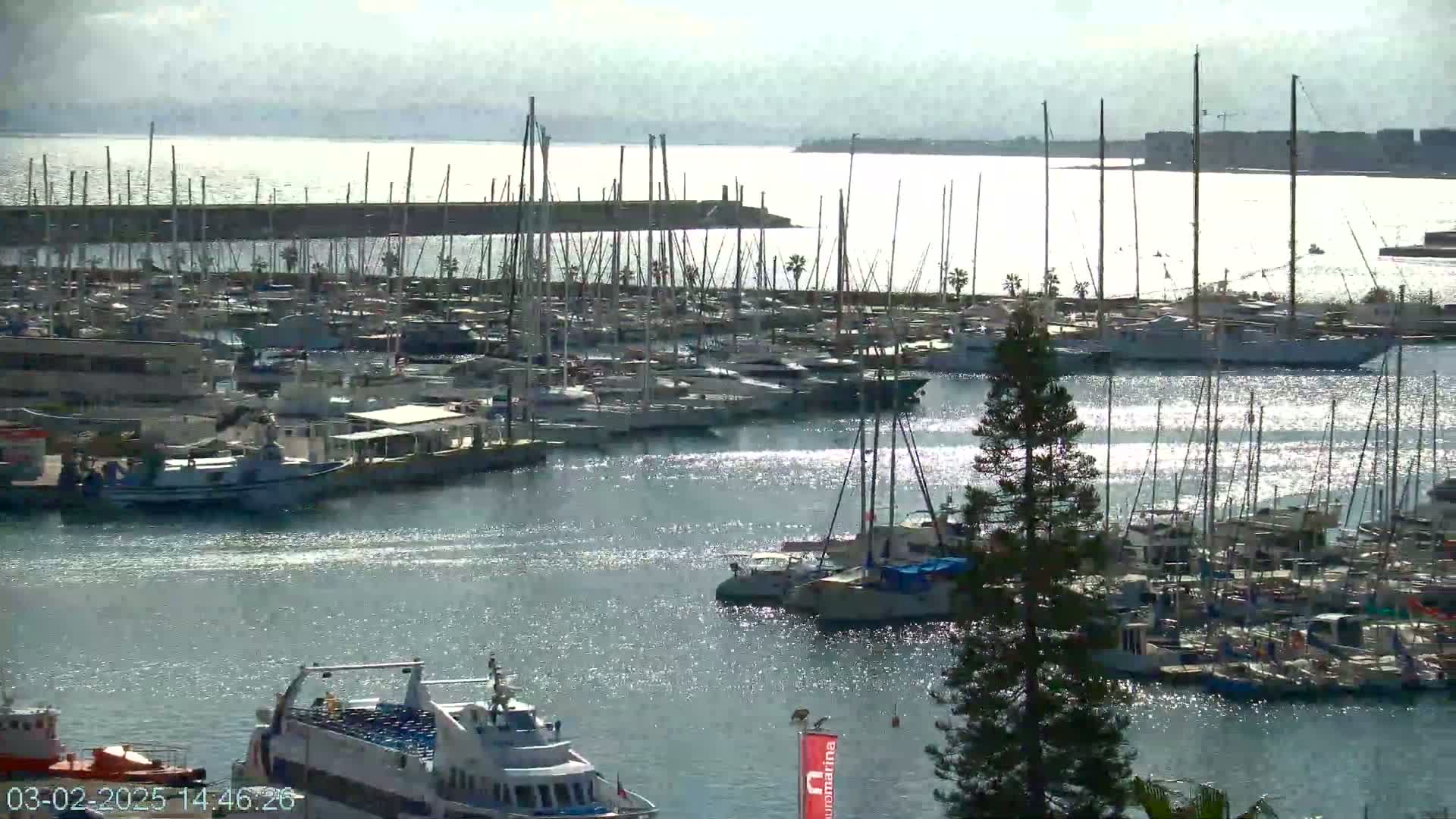 A marina filled with numerous sailboats and motorboats under a bright, mostly sunny sky, with calm water sparkling in the sunlight.