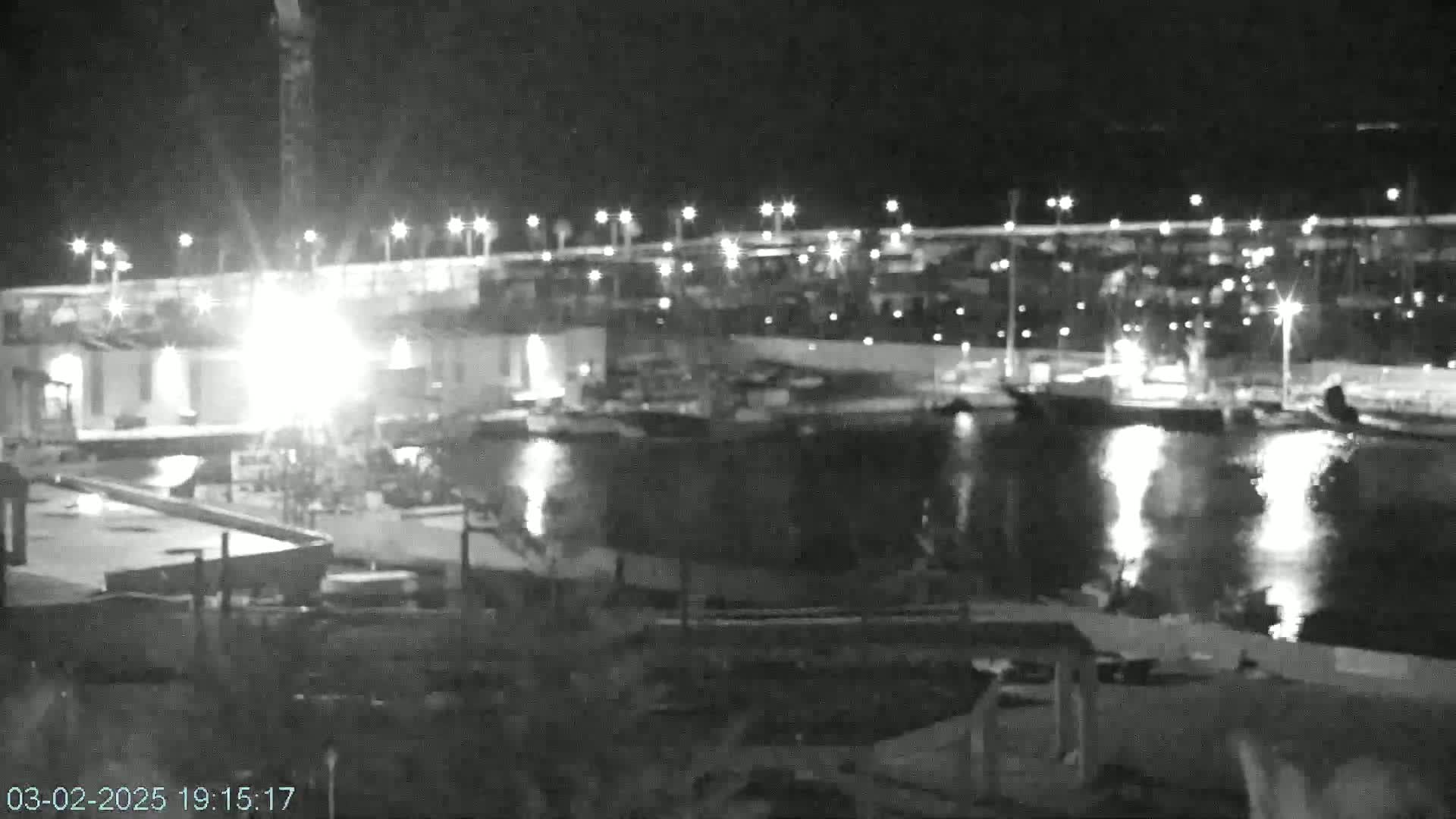 A nighttime, low-light view of a harbor with boats moored, lit by many streetlights and illuminated by bright lights near a construction site on the shore.