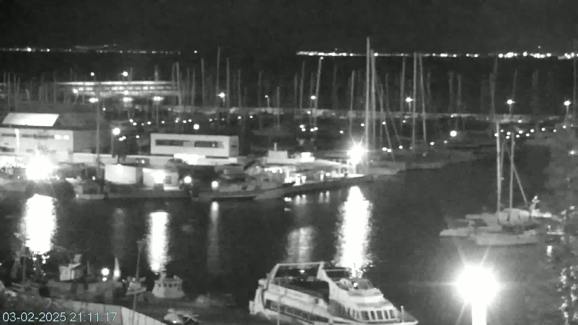 A nighttime, black-and-white view of a harbor filled with boats and buildings, reflecting lights on the calm water.