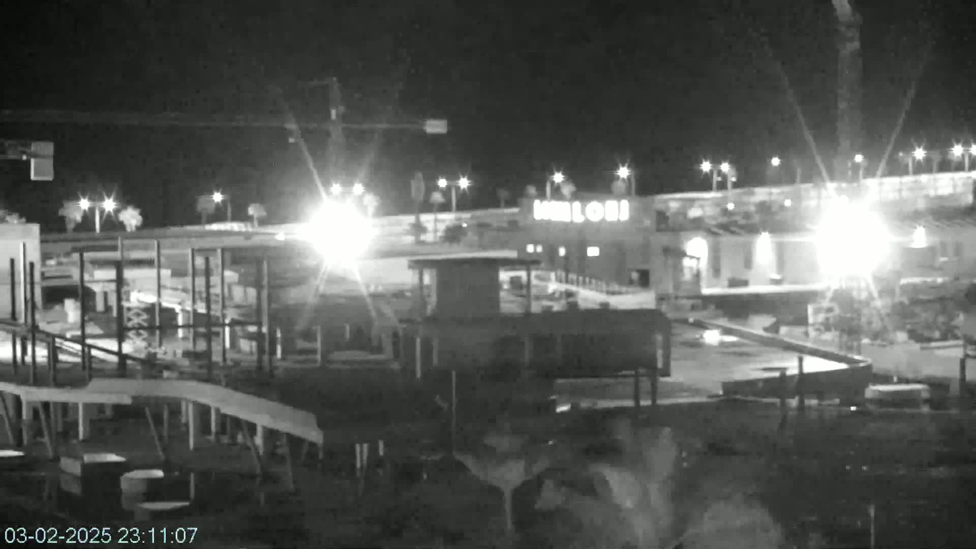 A nighttime, low-light grayscale image shows a building with lights on, a construction crane, and other structures in what appears to be a parking lot or industrial area.