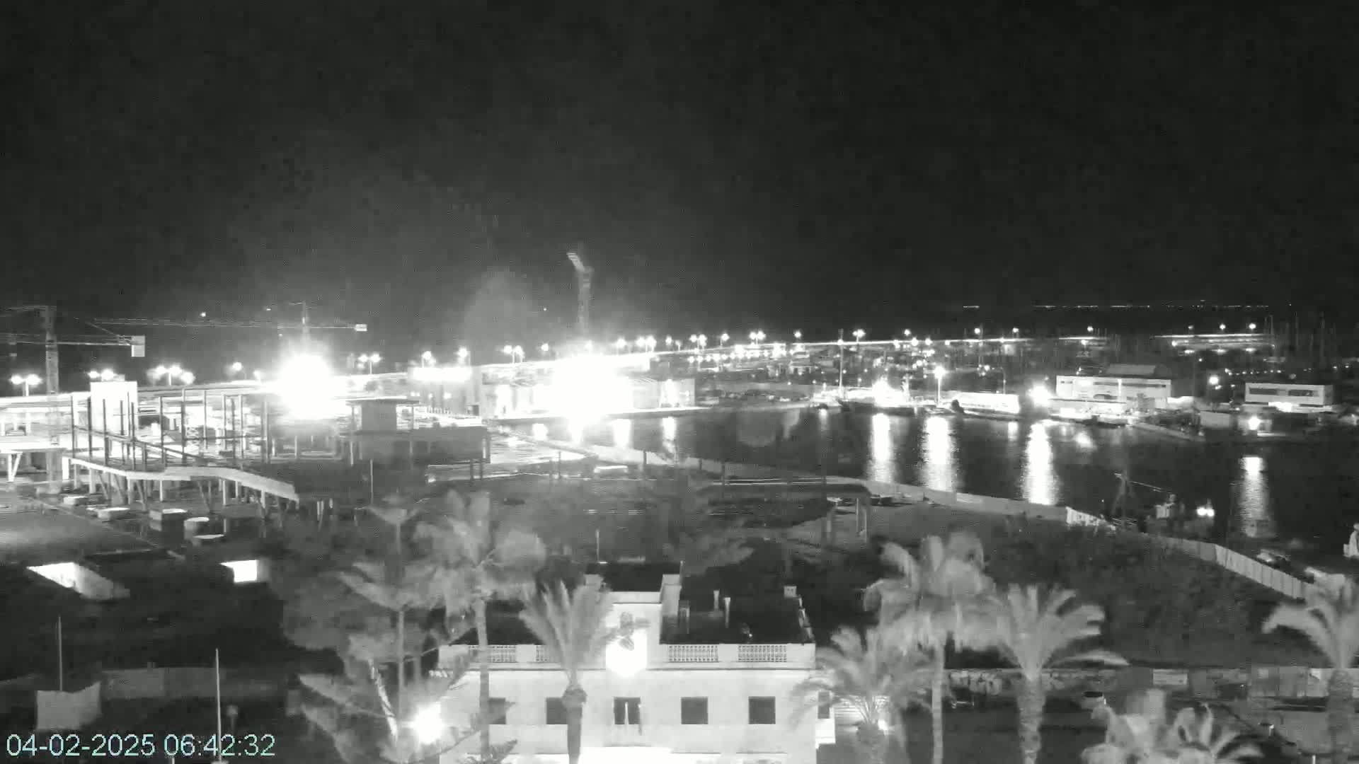 A nighttime view of a harbor with construction work in progress near palm trees and buildings under a clear sky.