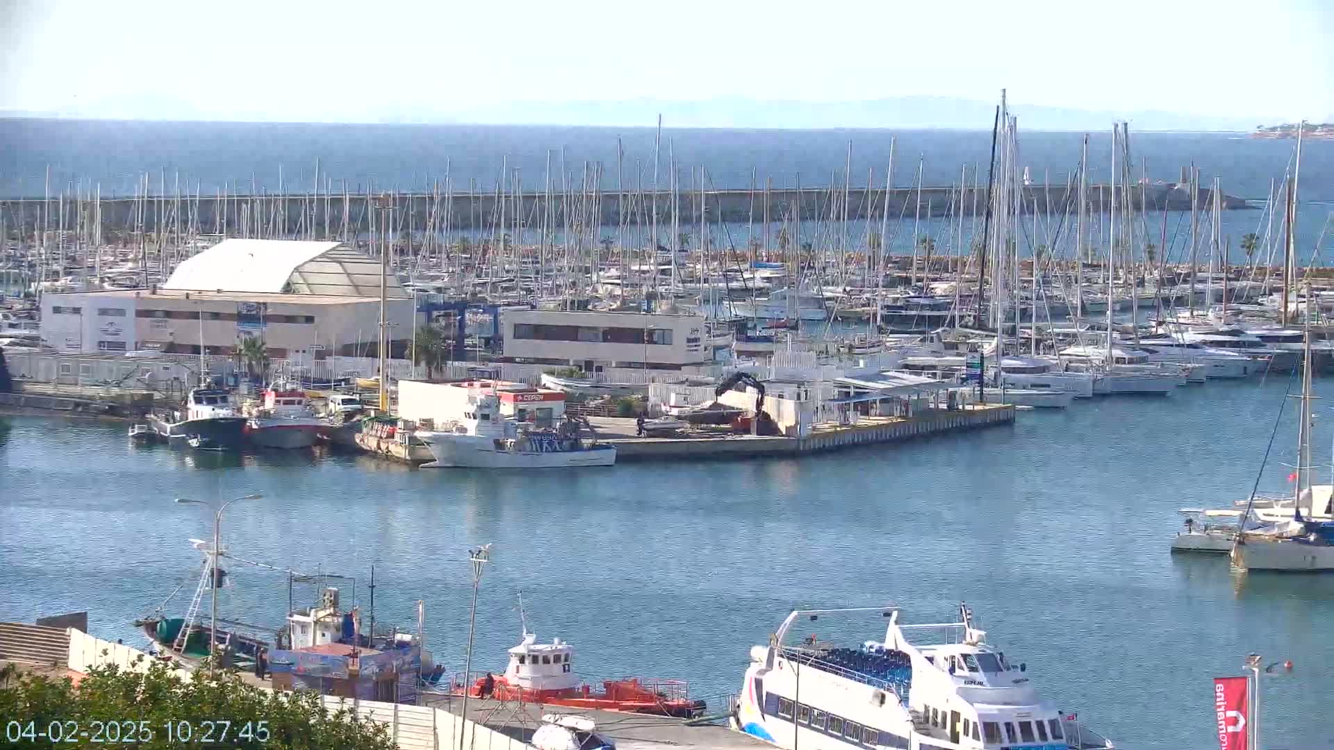 A marina filled with numerous sailboats and motorboats, under a clear sunny sky, is situated next to calm blue water and a distant coastline.