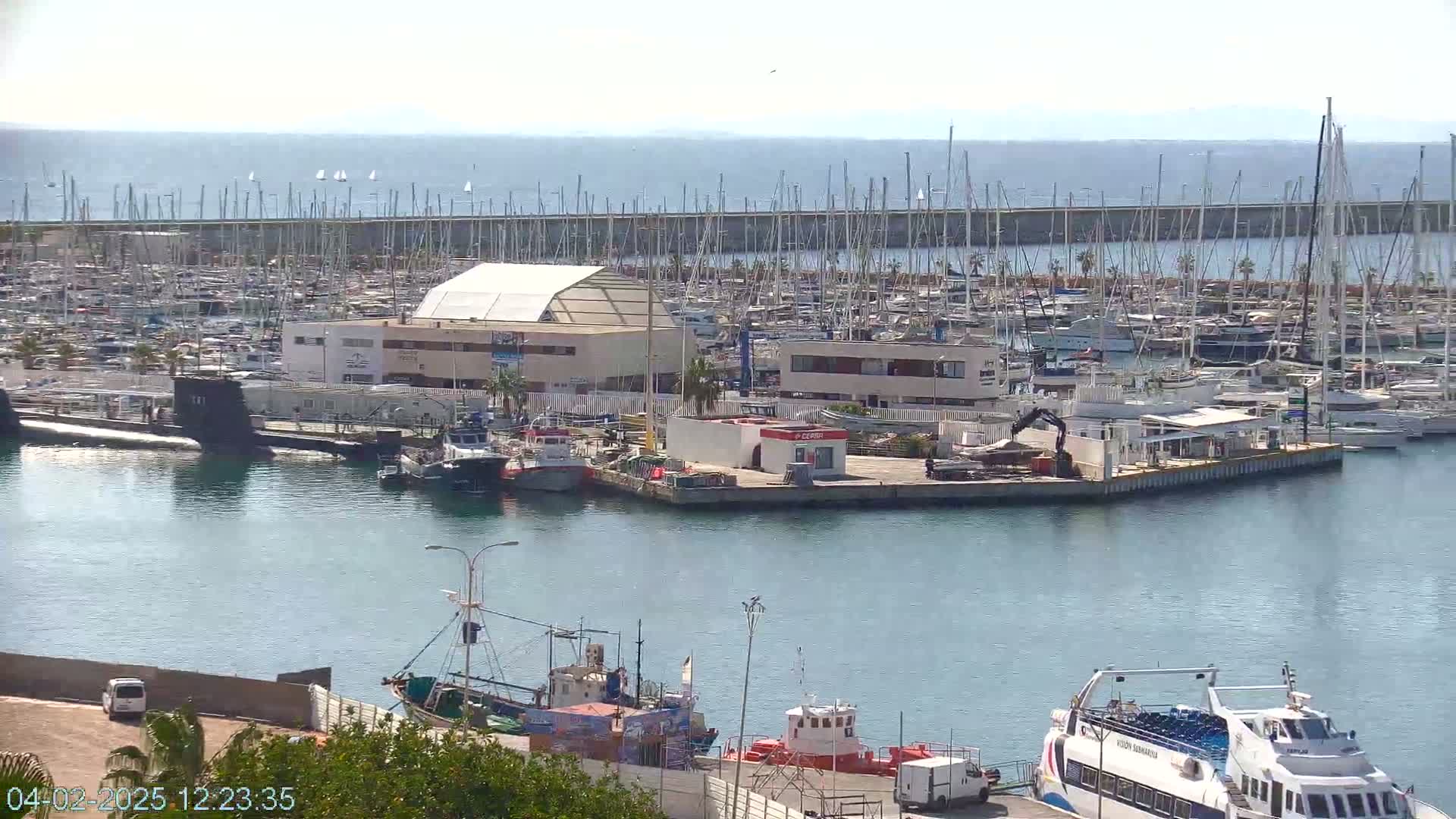 A marina filled with numerous boats and yachts under a sunny sky, with a submarine visible in the harbor.