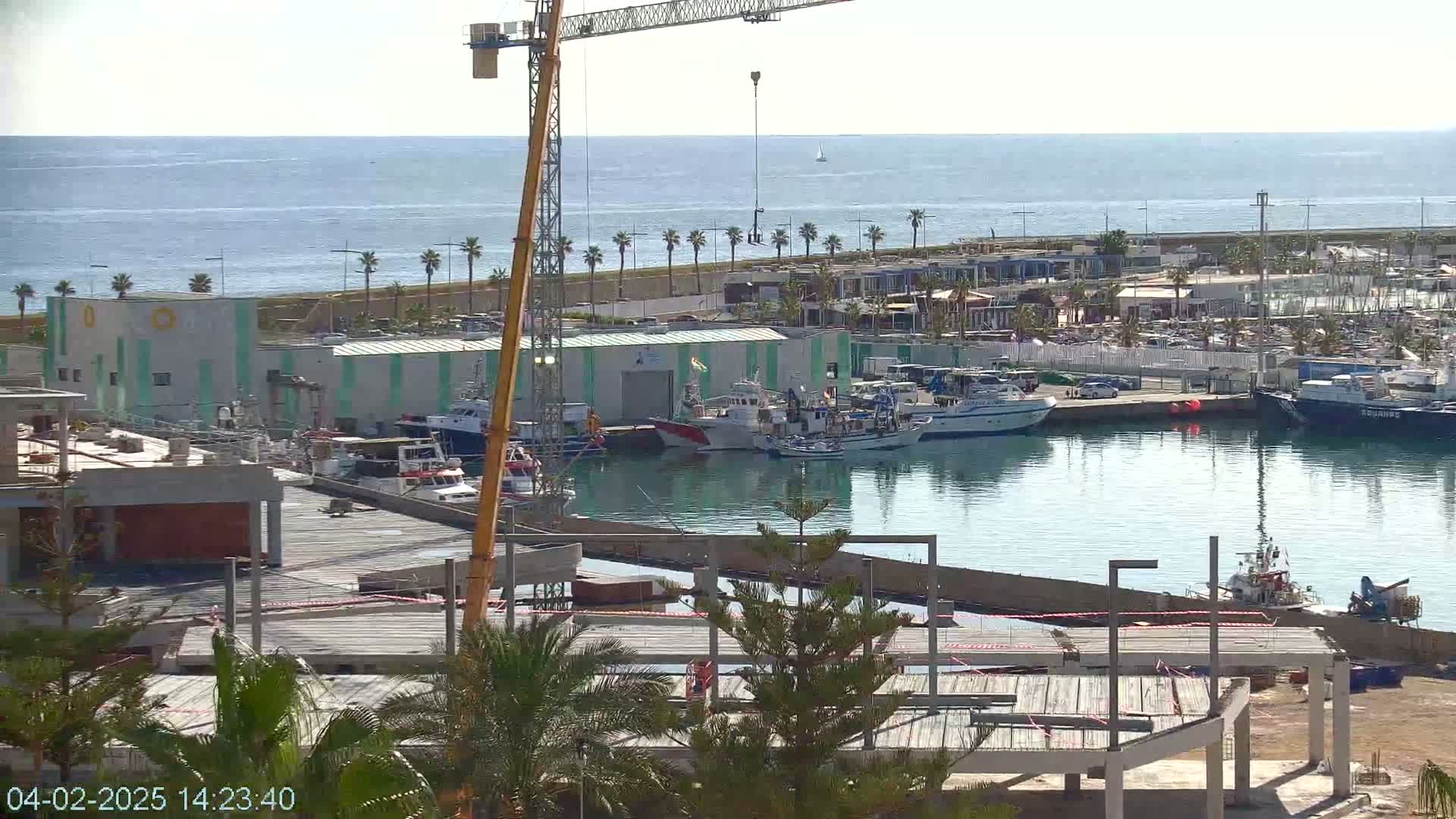 A harbor with numerous boats docked, a construction site in the foreground, and a calm ocean in the background under a clear sky.