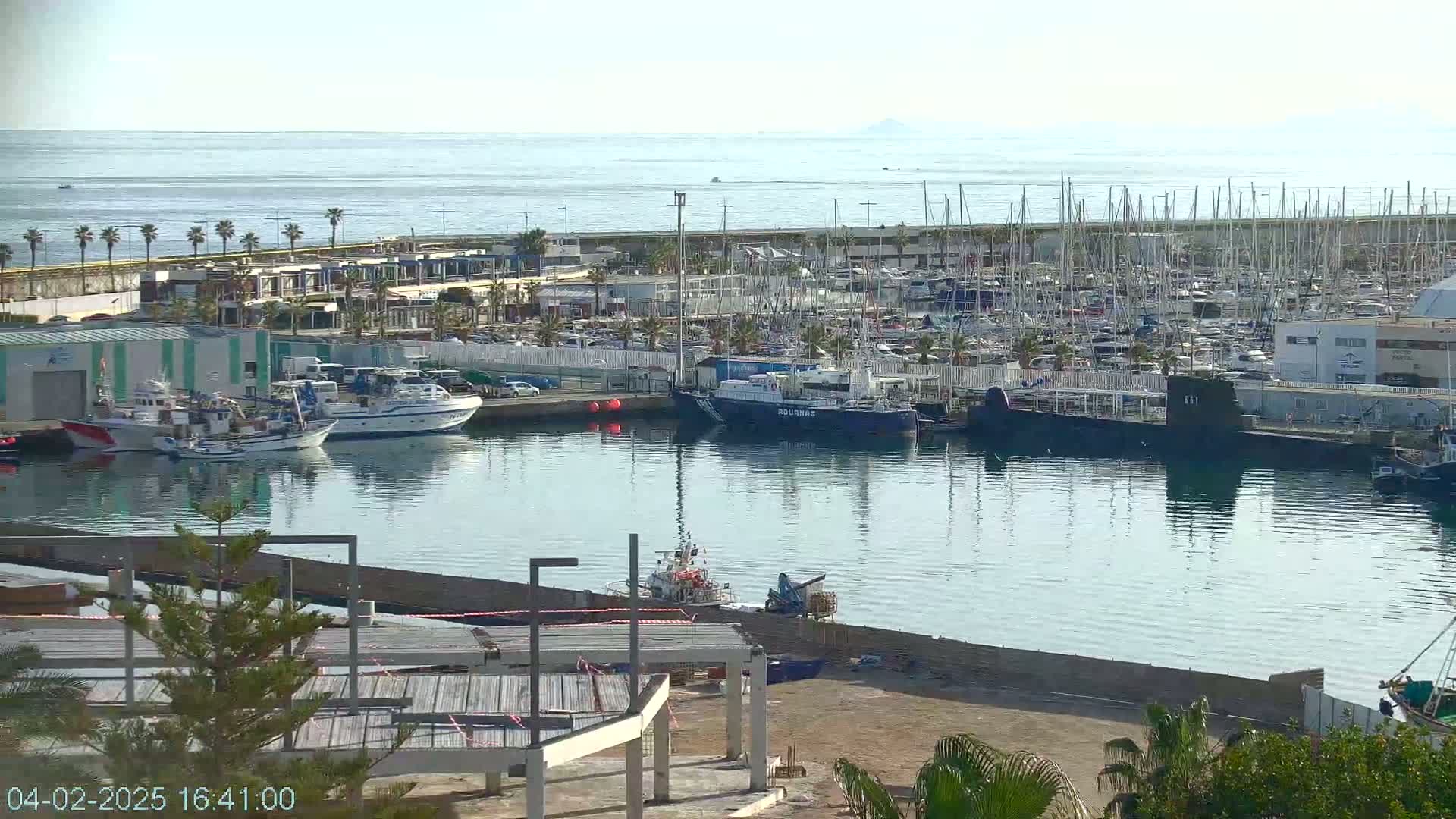 A calm harbor filled with numerous boats and yachts under a clear sky, with a distant view of the sea and land.