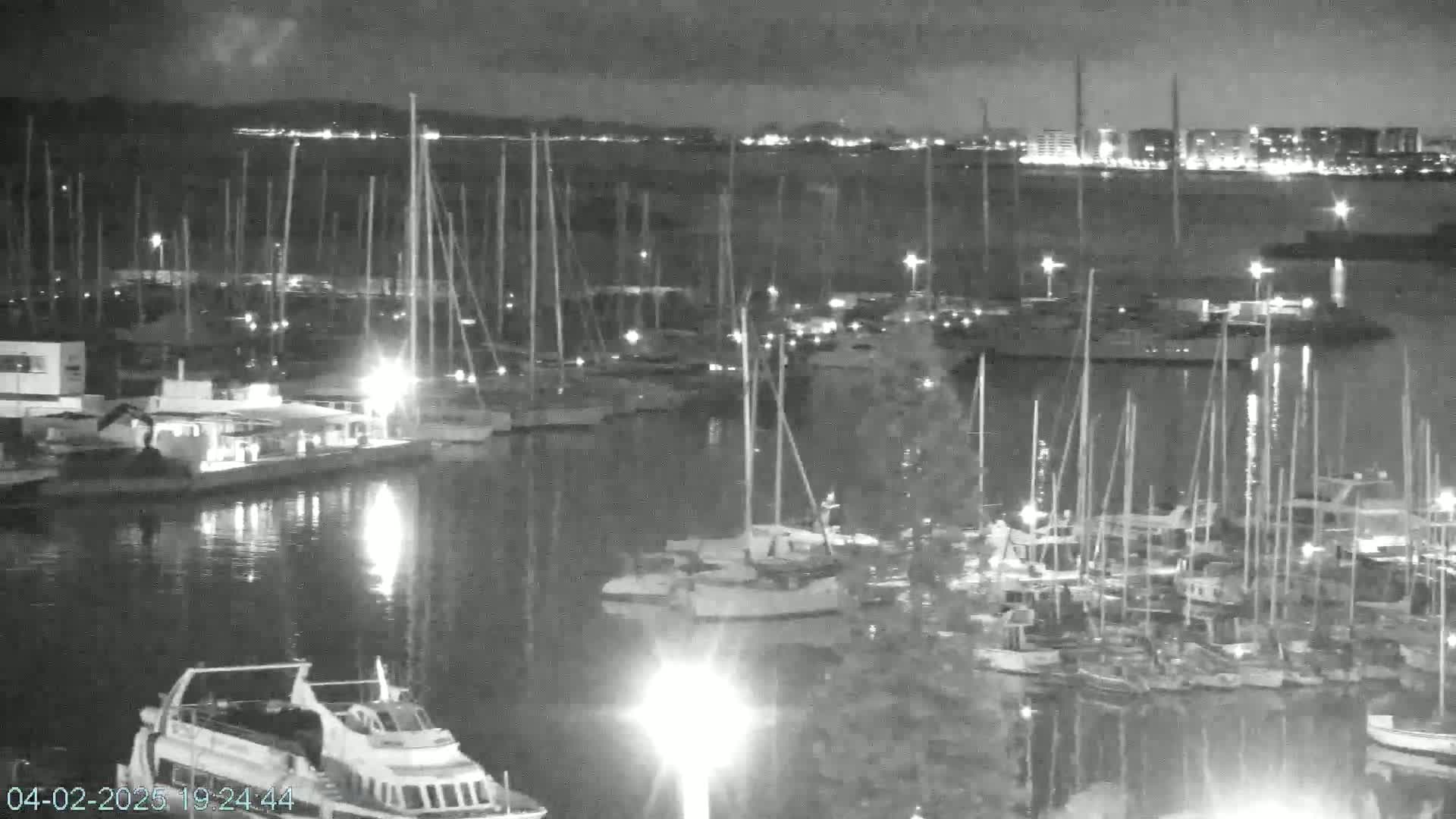 A nighttime view of a marina filled with numerous sailboats and motorboats, illuminated by lights, with a city skyline visible in the background under a dark, possibly cloudy sky.