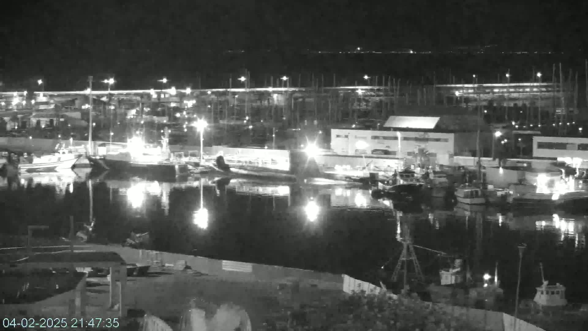 A nighttime view of a harbor filled with numerous boats and buildings, reflecting lights on calm water.