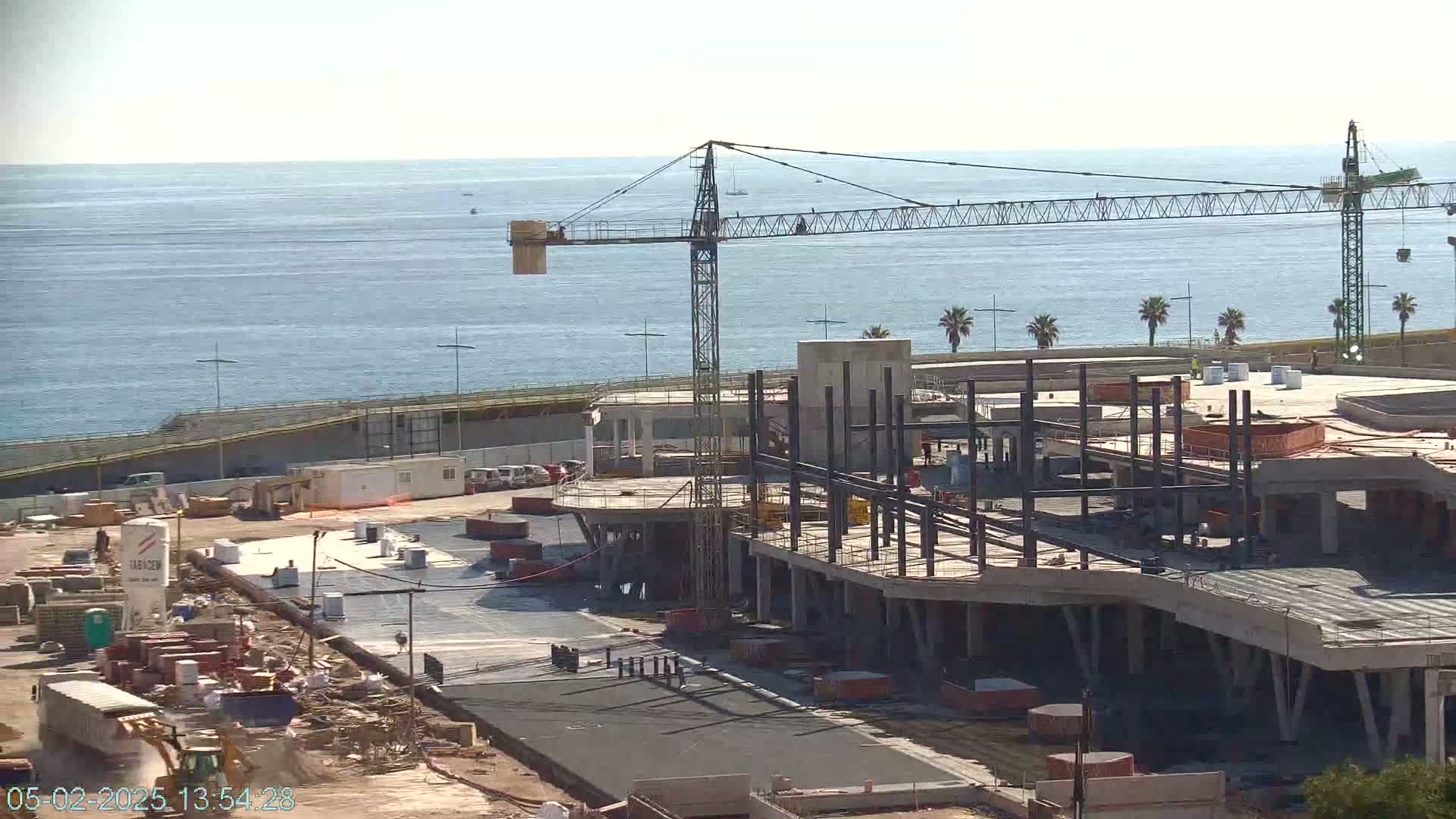 A large construction site near a calm ocean under a clear sky shows a partially built structure with a crane and various pieces of construction equipment.