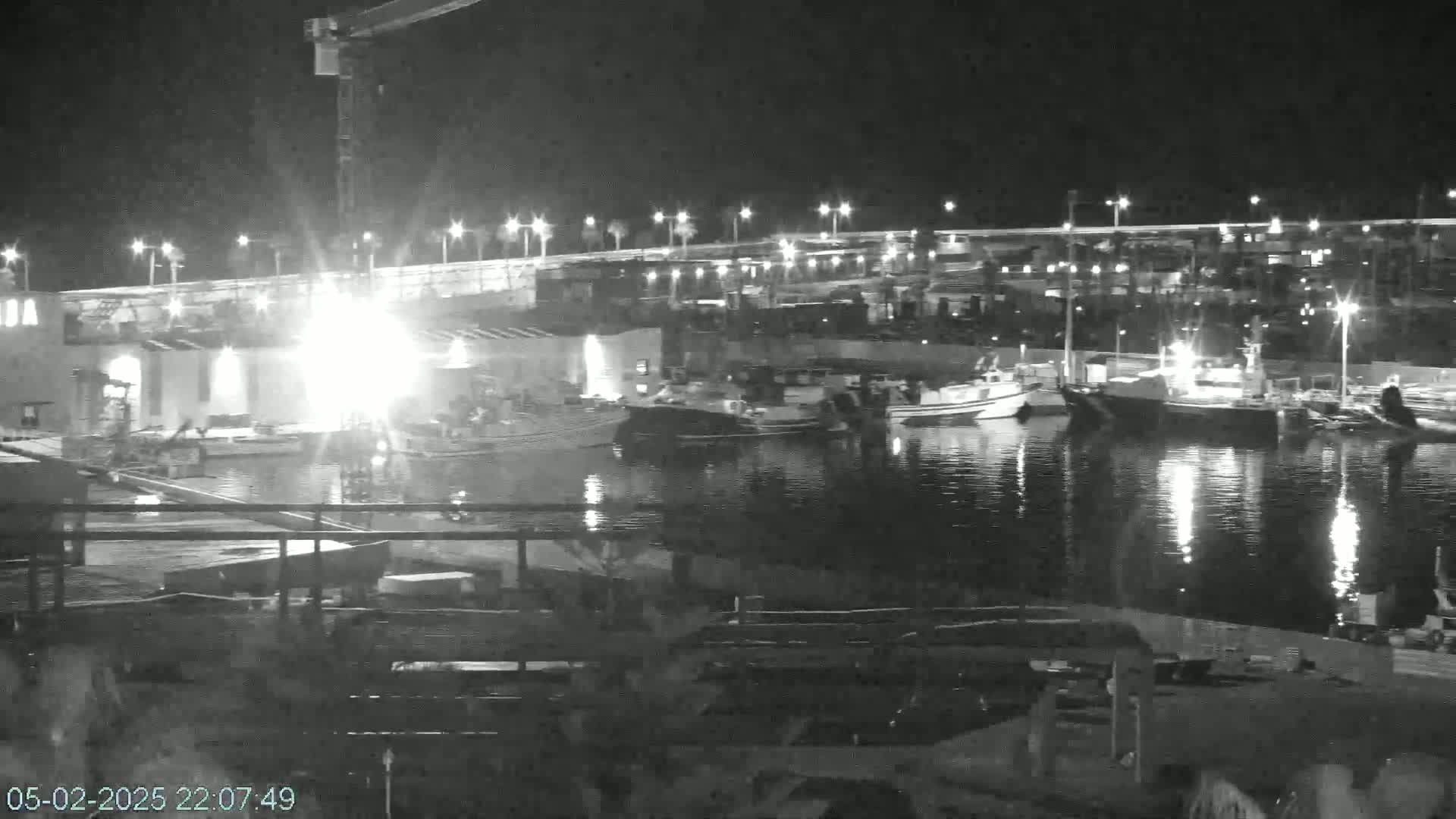 A nighttime view of a harbor with several boats docked, a construction crane, and bright lights illuminating the scene.