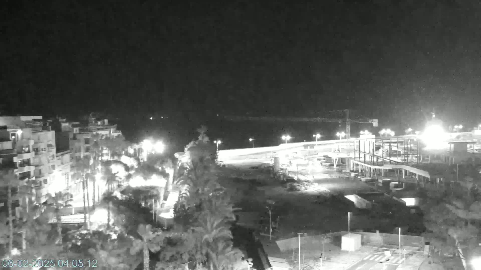 A nighttime, black-and-white view of a coastal area showing buildings, palm trees, and a construction site illuminated by artificial lights.