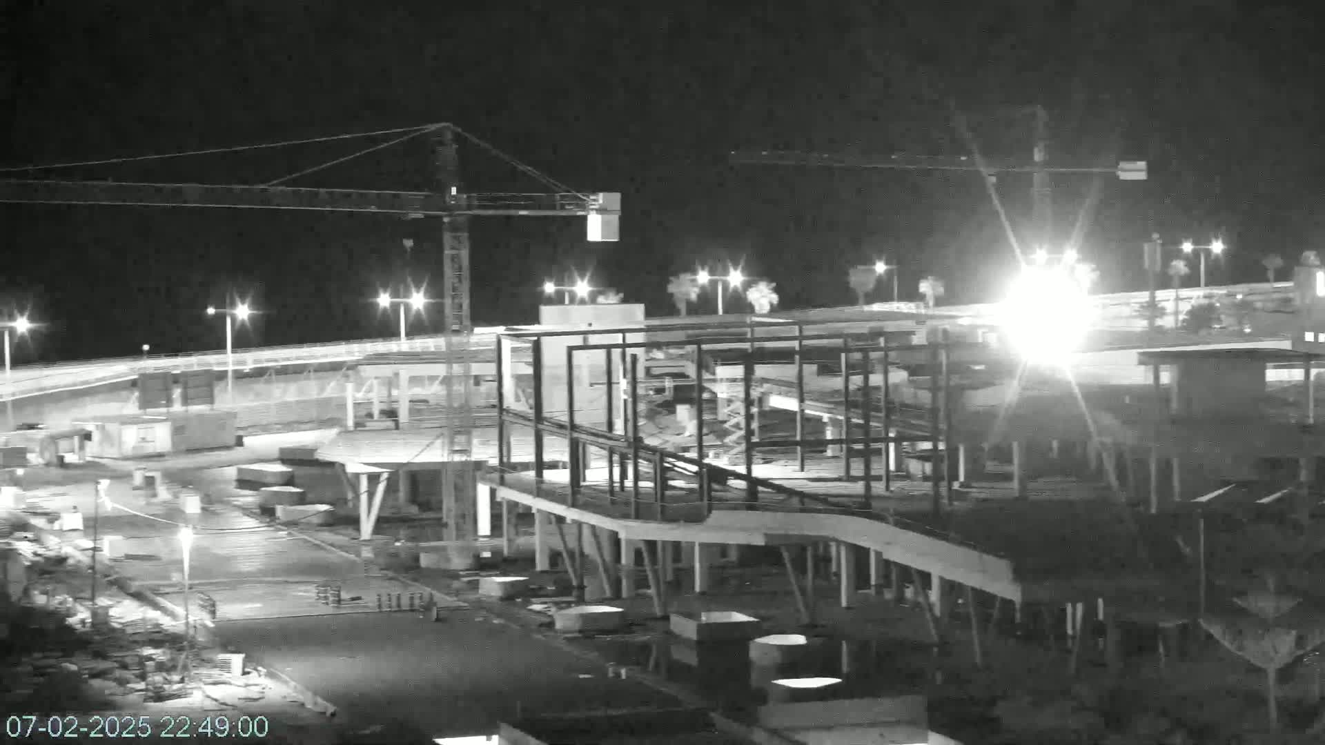 Under a dark night sky, a large building under construction is illuminated by artificial lights, with construction cranes visible in the background.