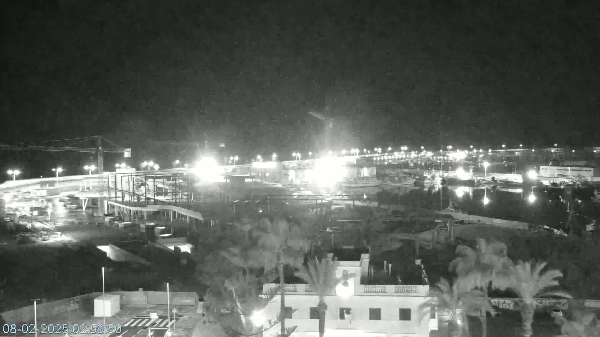 A nighttime, overhead view of a construction site with lights illuminating the area, adjacent to a palm tree-lined waterfront.