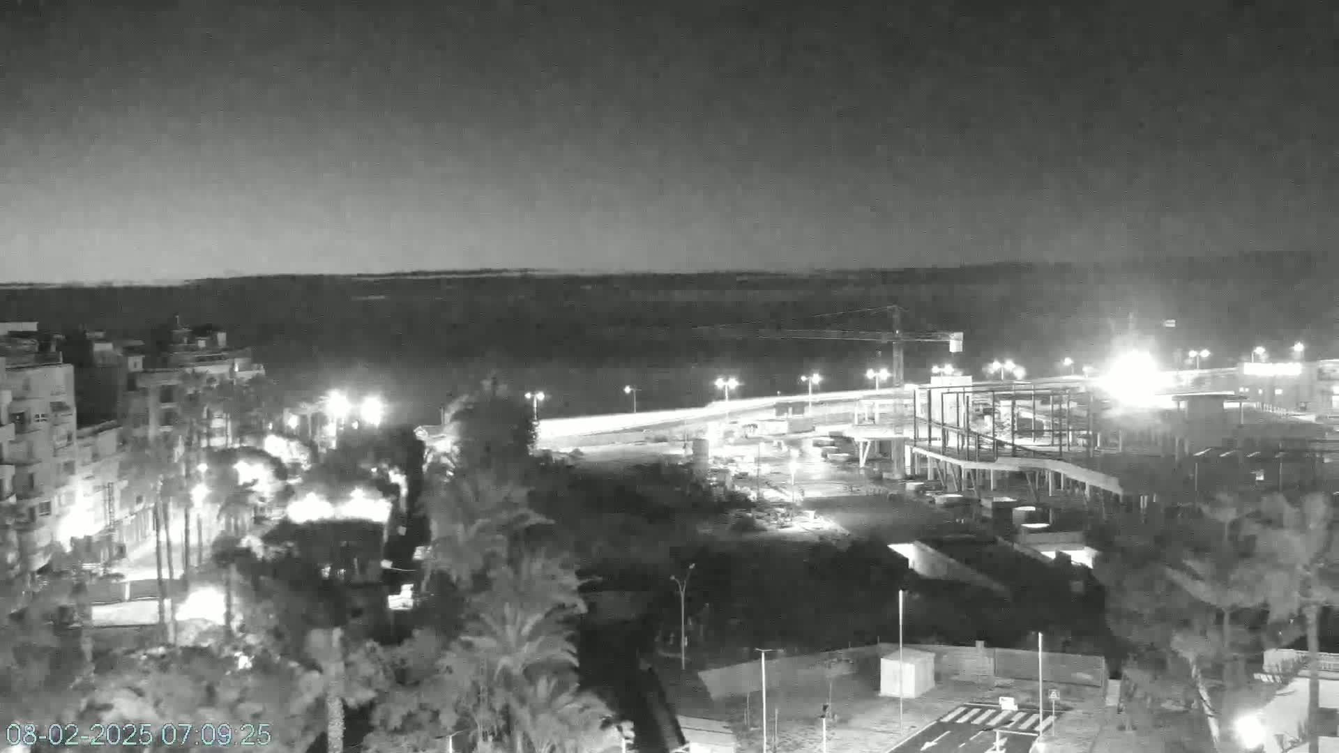 A nighttime, grayscale view shows a coastal town with palm trees, buildings under construction, and a roadway lit by streetlights, under a dark, possibly cloudy sky.