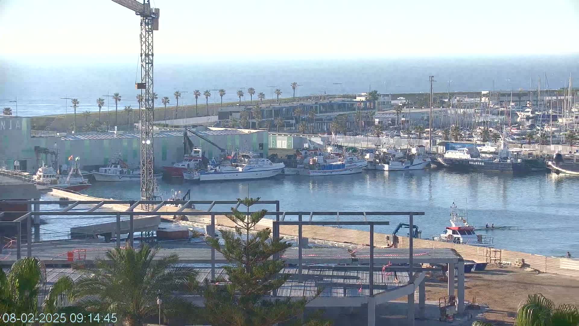 A harbor filled with numerous boats and ships under a clear sky, with a construction site in the foreground and a crane visible.