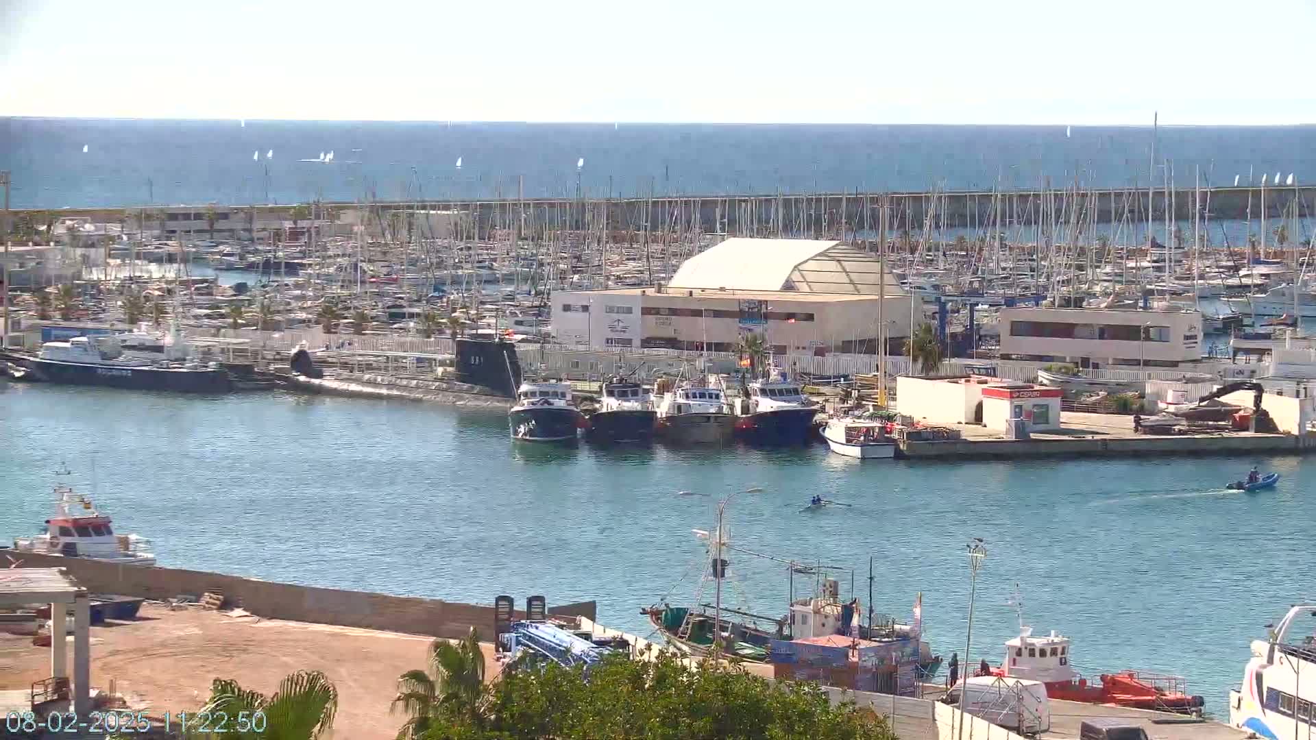 A marina filled with numerous boats and yachts under a sunny sky, with a calm sea visible in the background.