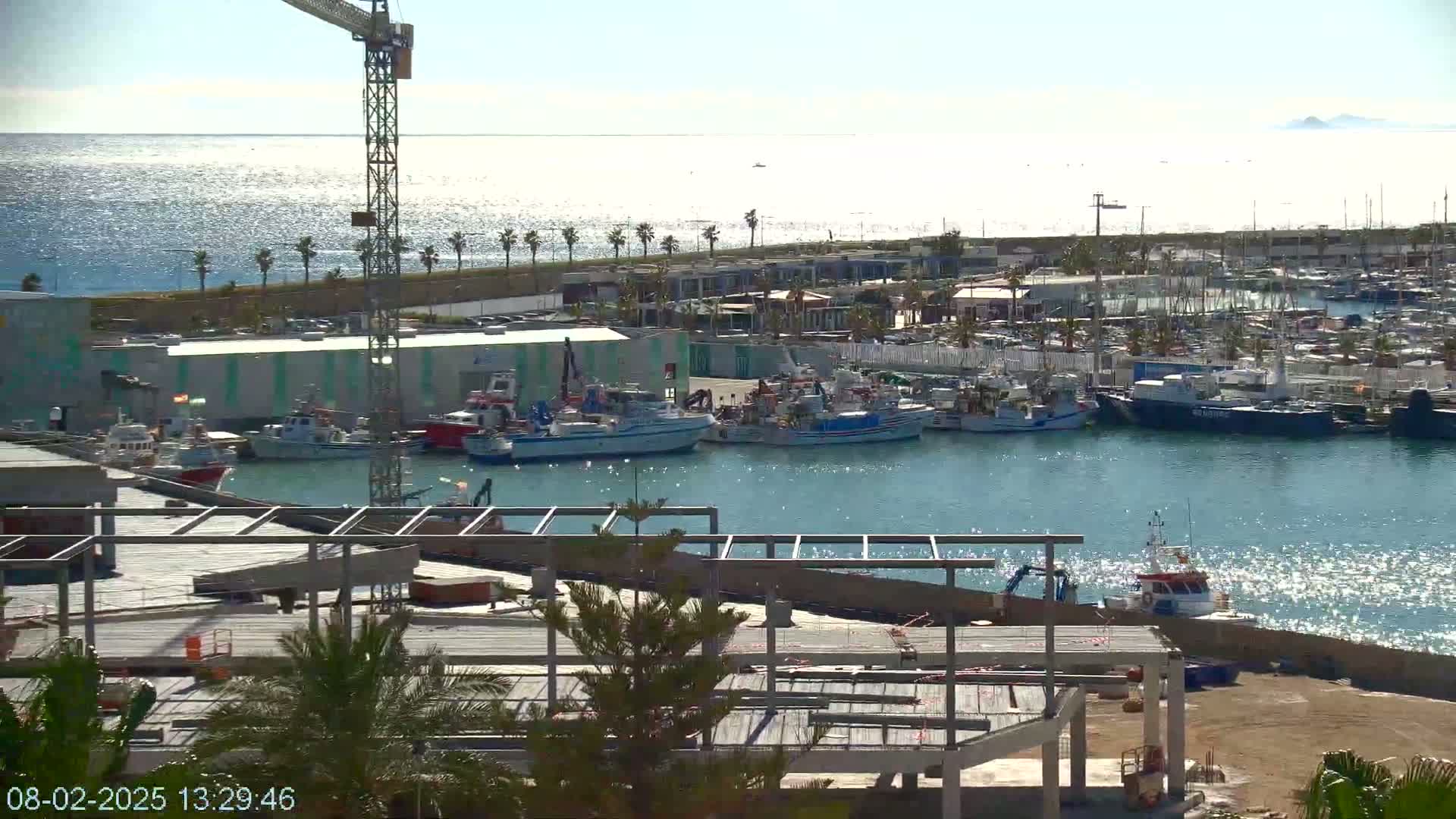 A harbor filled with numerous boats, a construction site, and a tall crane under sunny skies.