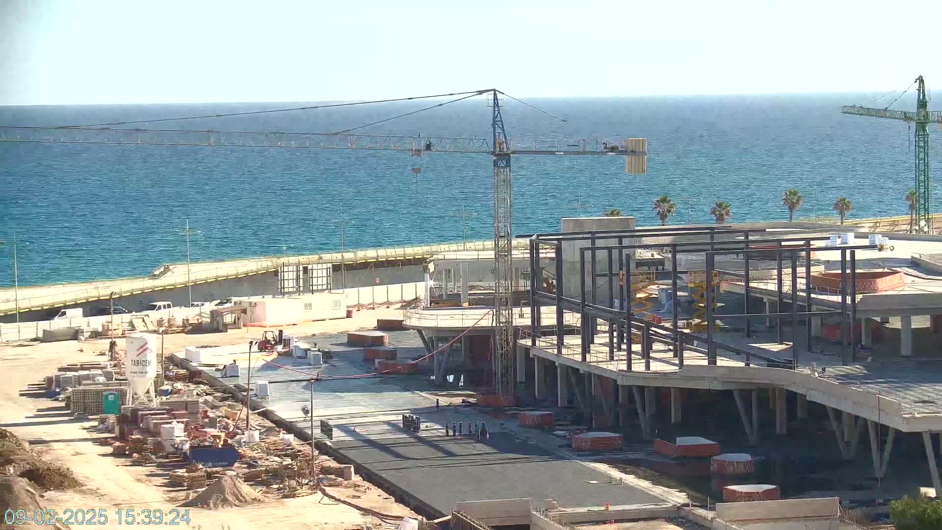 A seaside construction site shows a partially built structure with a crane and other construction equipment under a clear sky.