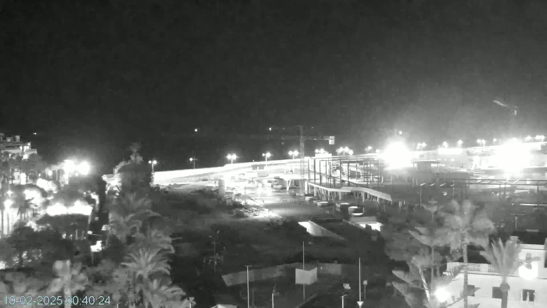 A nighttime, aerial view of a coastal construction site with palm trees and brightly lit buildings under a clear, dark sky.