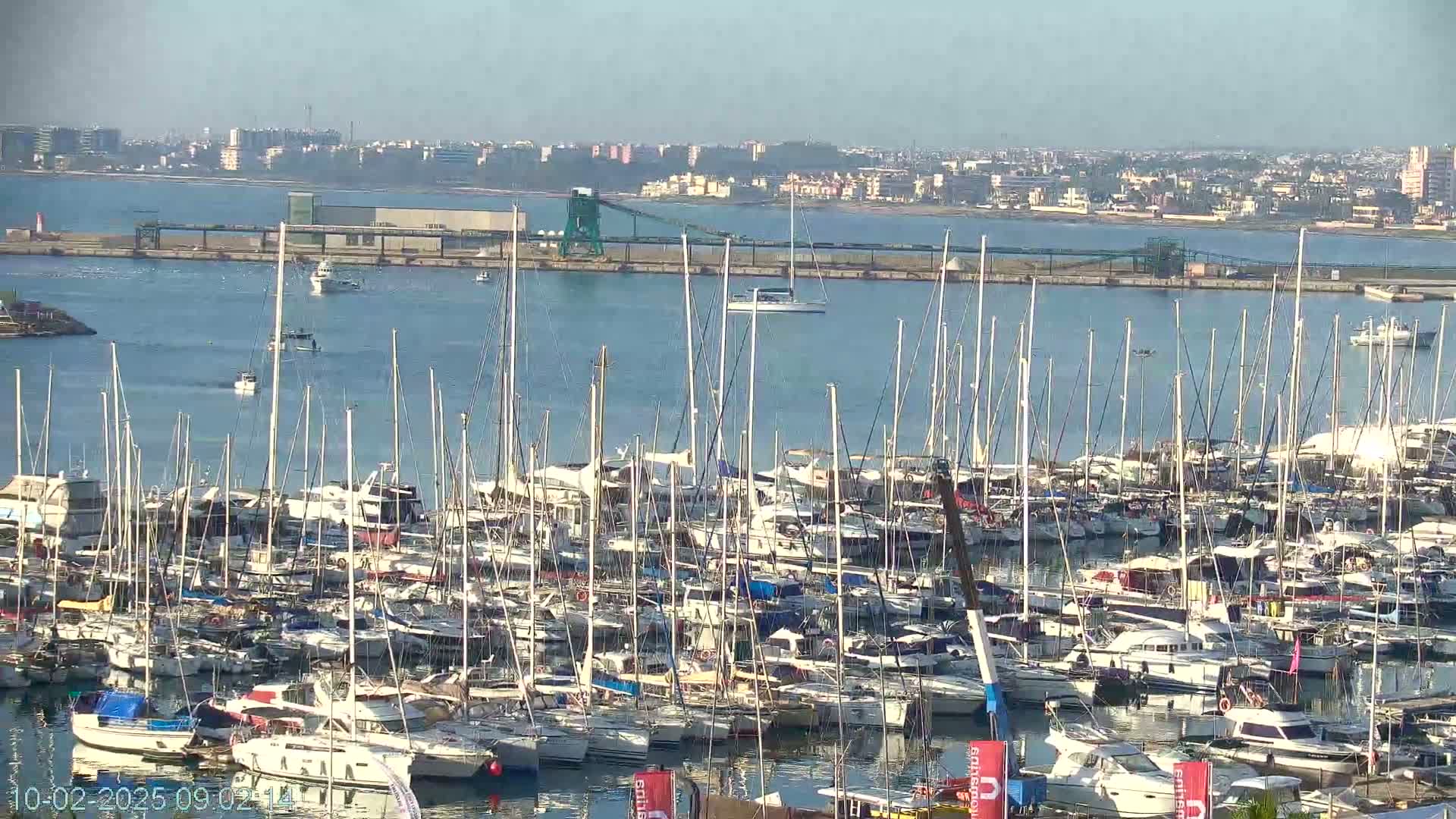 A large marina filled with numerous sailboats and motorboats is situated on calm water under a clear sky, with a distant cityscape visible in the background.