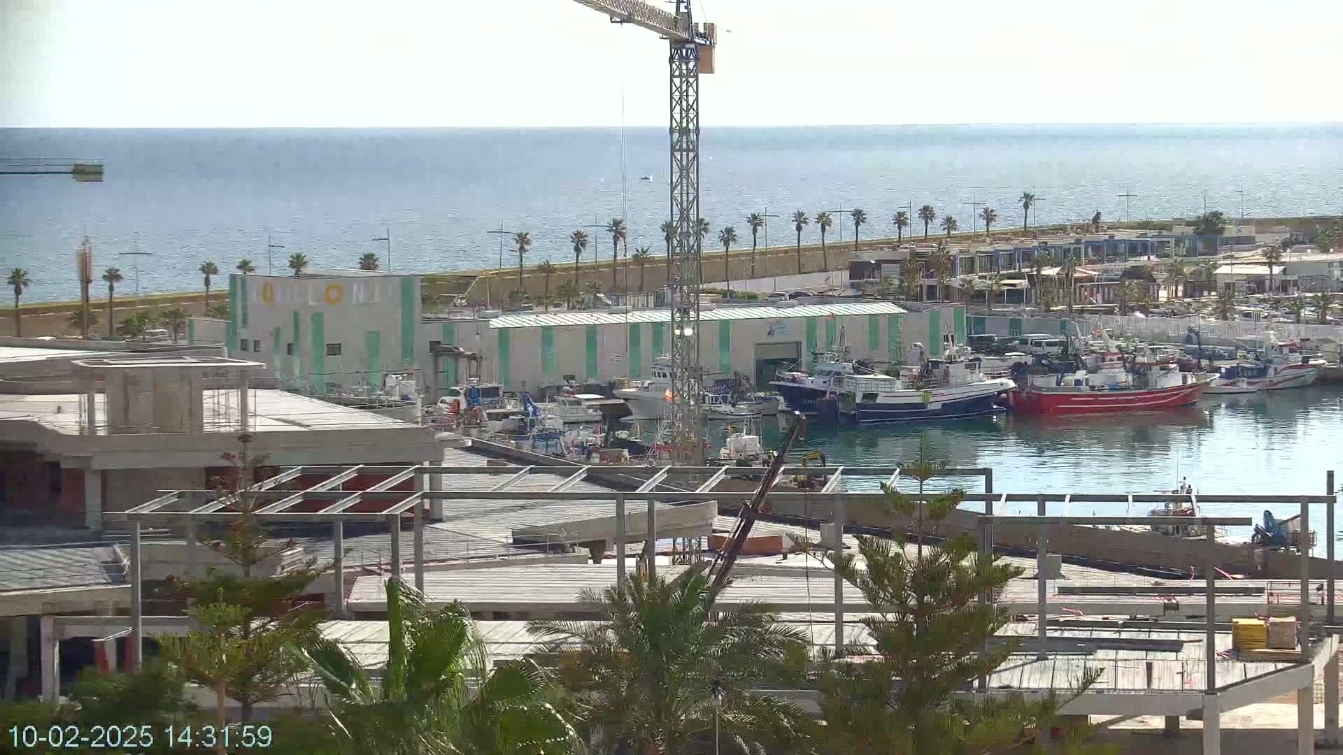 A harbor filled with fishing boats is seen from a distance, with a construction site in the foreground under a clear, sunny sky.