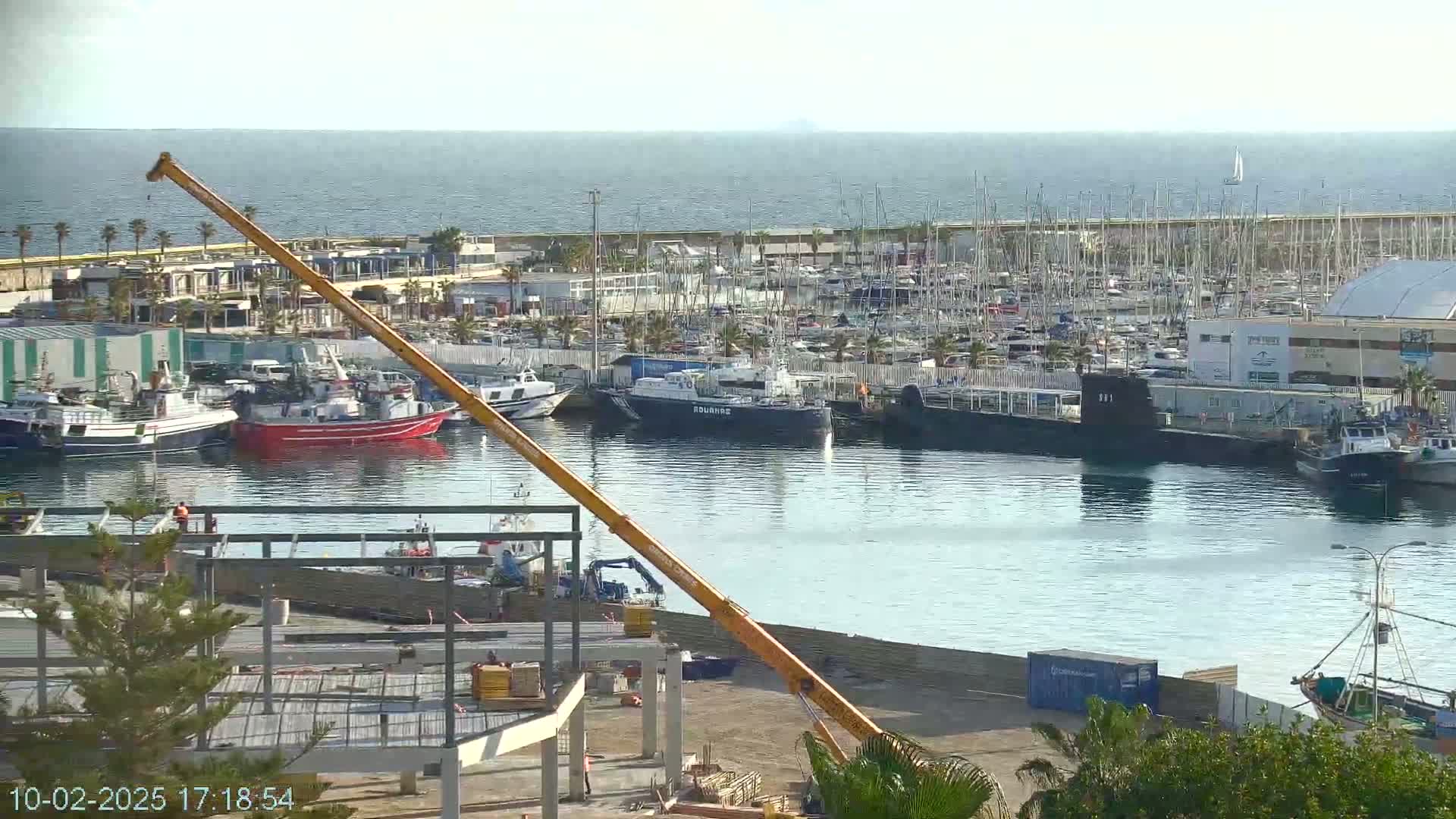 A large yellow crane overlooks a harbor filled with numerous boats and ships under a clear, sunny sky.