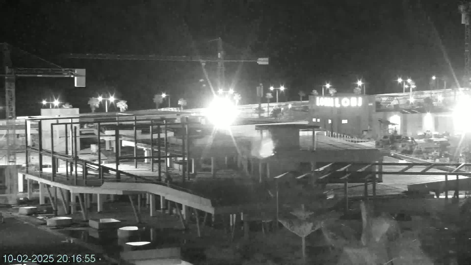 A nighttime, black and white image shows a building under construction with steel framework and supports visible against a background of lights and a partially completed structure.