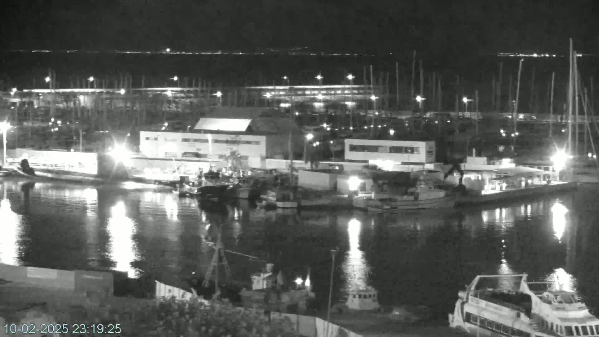 A nighttime, black and white view of a harbor filled with docked boats and illuminated buildings, reflecting in calm water under a dark sky.