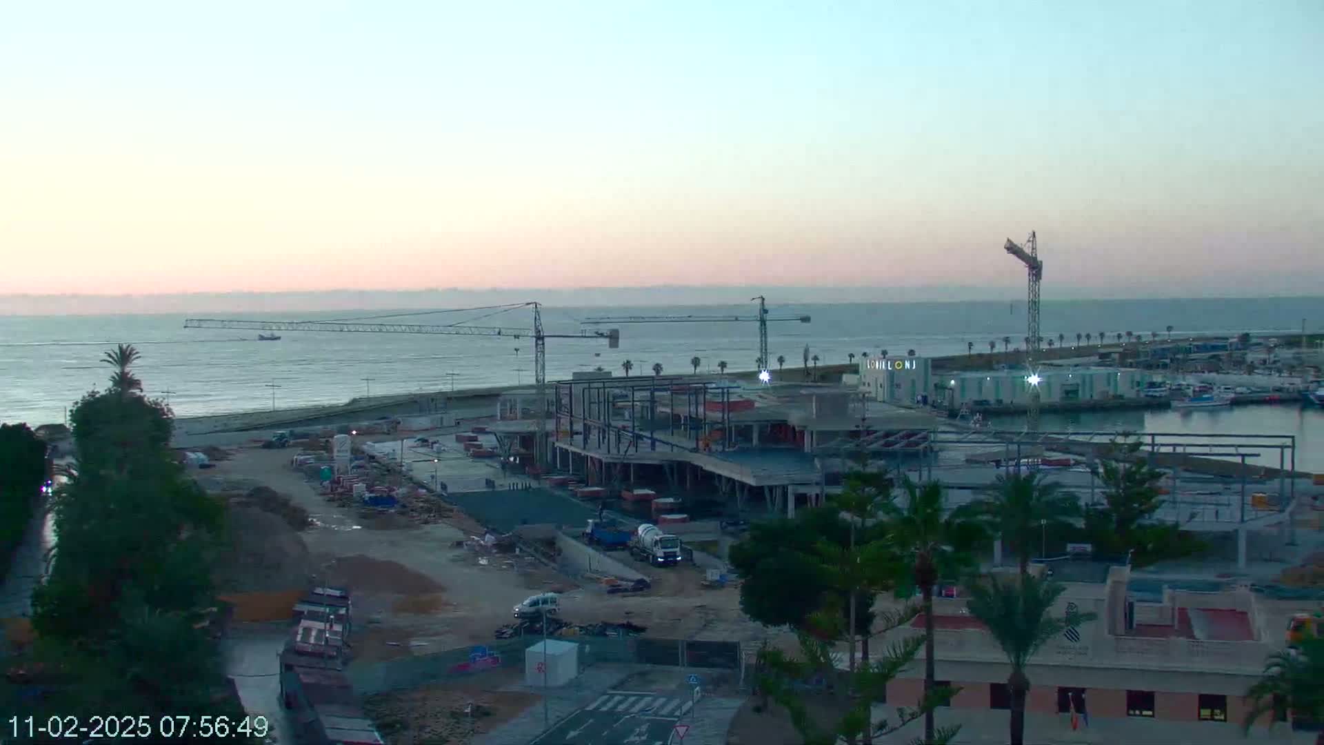 A coastal construction site with several cranes is visible at dawn, under a pale pink sky and calm sea.