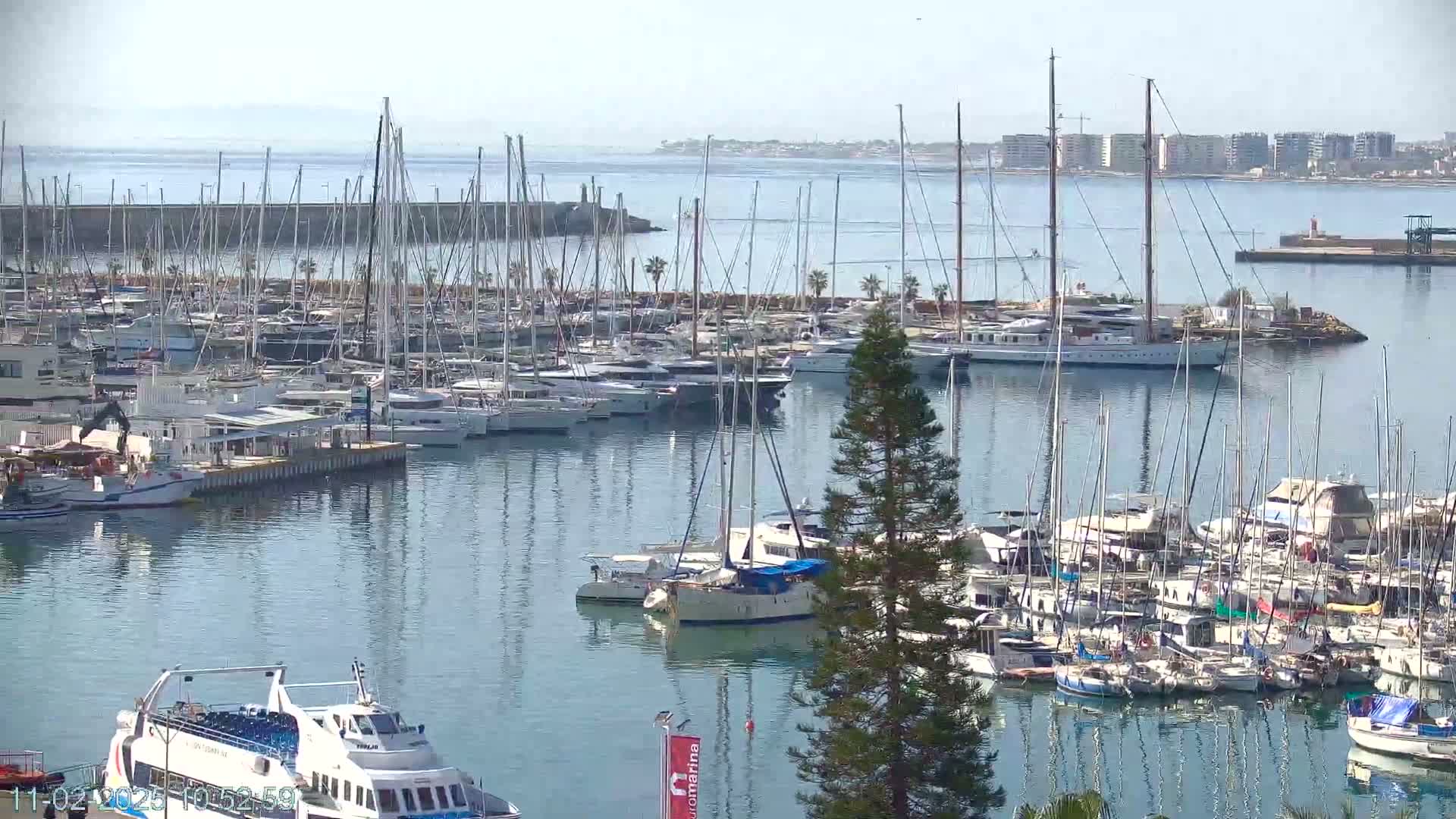 A large marina filled with numerous sailboats and motorboats, under a mostly clear sky, with a calm body of water extending to a distant shoreline.