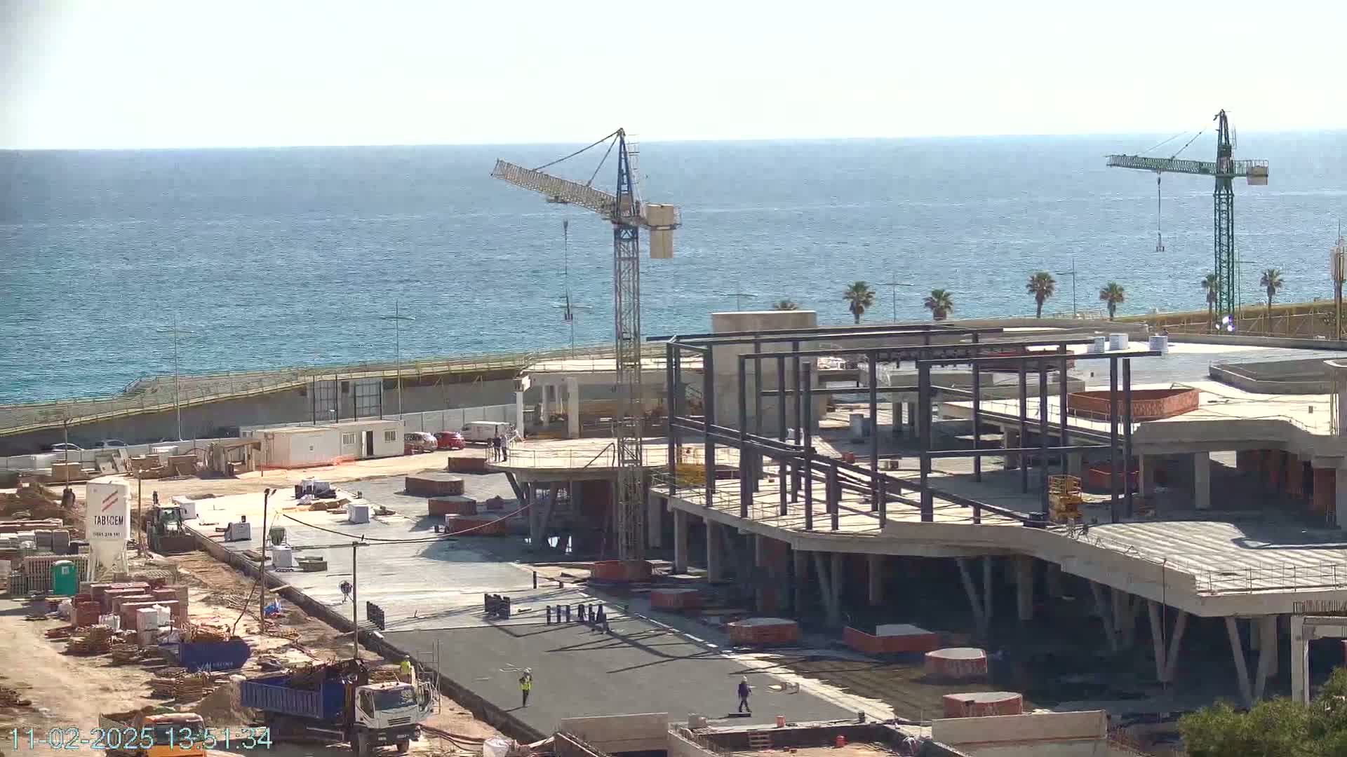 A coastal construction site with two cranes is under a clear sky, next to a calm ocean.